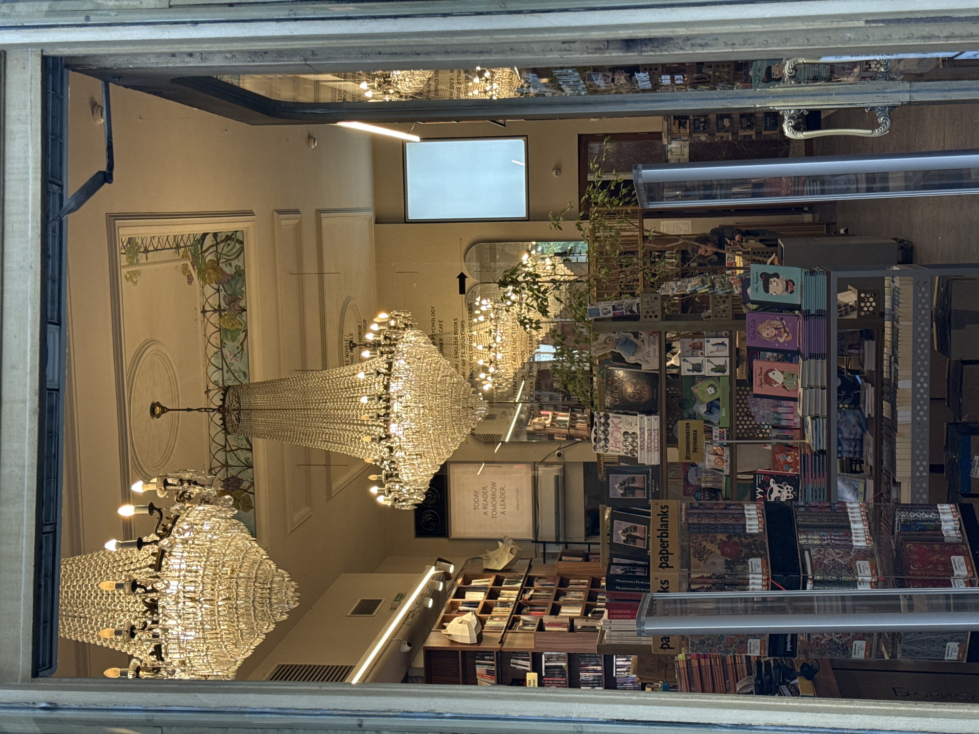 This indoor, wide-angle, high-angle shot captures the interior of a bookstore through a window in Belgrade, Serbia. The image appears to be reflected due to the angle of the window. In the foreground, there are rows of books with various covers and titles, including one titled "paperblanks". The shelves holding the books are metallic. In the middle ground, a sign reads "TODAY A READER, TOMORROW A LEADER." Behind the sign are bookshelves filled with books. In the background, there are ornate chandeliers hanging from the ceiling. The walls are off-white and feature decorative molding and artwork. A large, rectangular light source is visible on the ceiling. The bookstore appears to be well-lit and organized. The overall scene suggests a sophisticated and cultural environment.