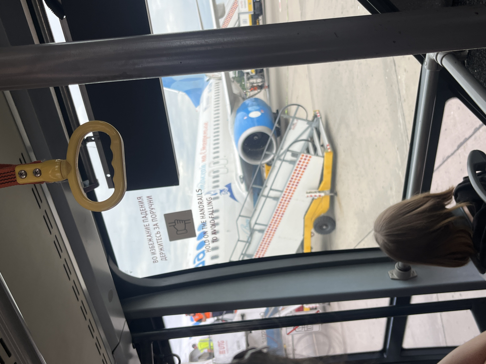 This media file captures a view from inside a bus or shuttle, looking out towards an airplane parked at an airport. The primary subjects are the airplane, a set of mobile stairs leading to the aircraft, and a glimpse of a child with brown hair inside the bus. The scene is set on an airport tarmac, with the ground appearing to be concrete or asphalt.
The airplane is white with blue accents and has "Nor" visible on its fuselage, suggesting it's a commercial passenger jet. The mobile stairs are yellow and white, with red checkered markings on the steps. Text is visible on the bus window, including "ВО ИЗБЕЖАНИЕ ПАДЕНИЯ ДЕРЖИТЕСЬ ЗА ПОРУЧНИ" (To avoid falling, hold onto the handrails) in Russian, and "HOLD ON THE HANDRAILS TO AVOID FALLING" in English, accompanied by a pictogram of a hand on a railing. Other visible text on the bus includes "EUTG" and "MADP".
The lighting suggests it is daytime, likely under a cloudy sky, as there are no strong shadows and the overall light is diffused. The presence of the airplane, stairs, and what appears to be ground crew in the background indicates an active airport environment, possibly during boarding or disembarking. The focus on the warning labels on the window and the obscured view of the child inside create a sense of being in transit within the airport.