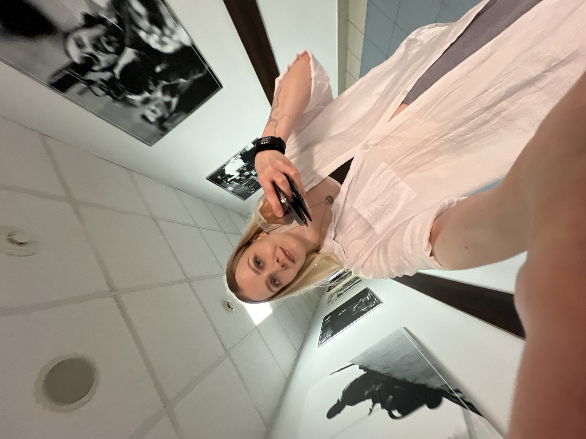 A woman is taking a selfie in an indoor space with a white tiled ceiling with circular light fixtures and black and white artwork lining the walls. She is holding a pair of tweezers or pliers in her left hand. She has blonde hair and is wearing a white shirt over a gray top. The location appears to be an art gallery or similar space, possibly in Khimki, Russia based on context not visually present.