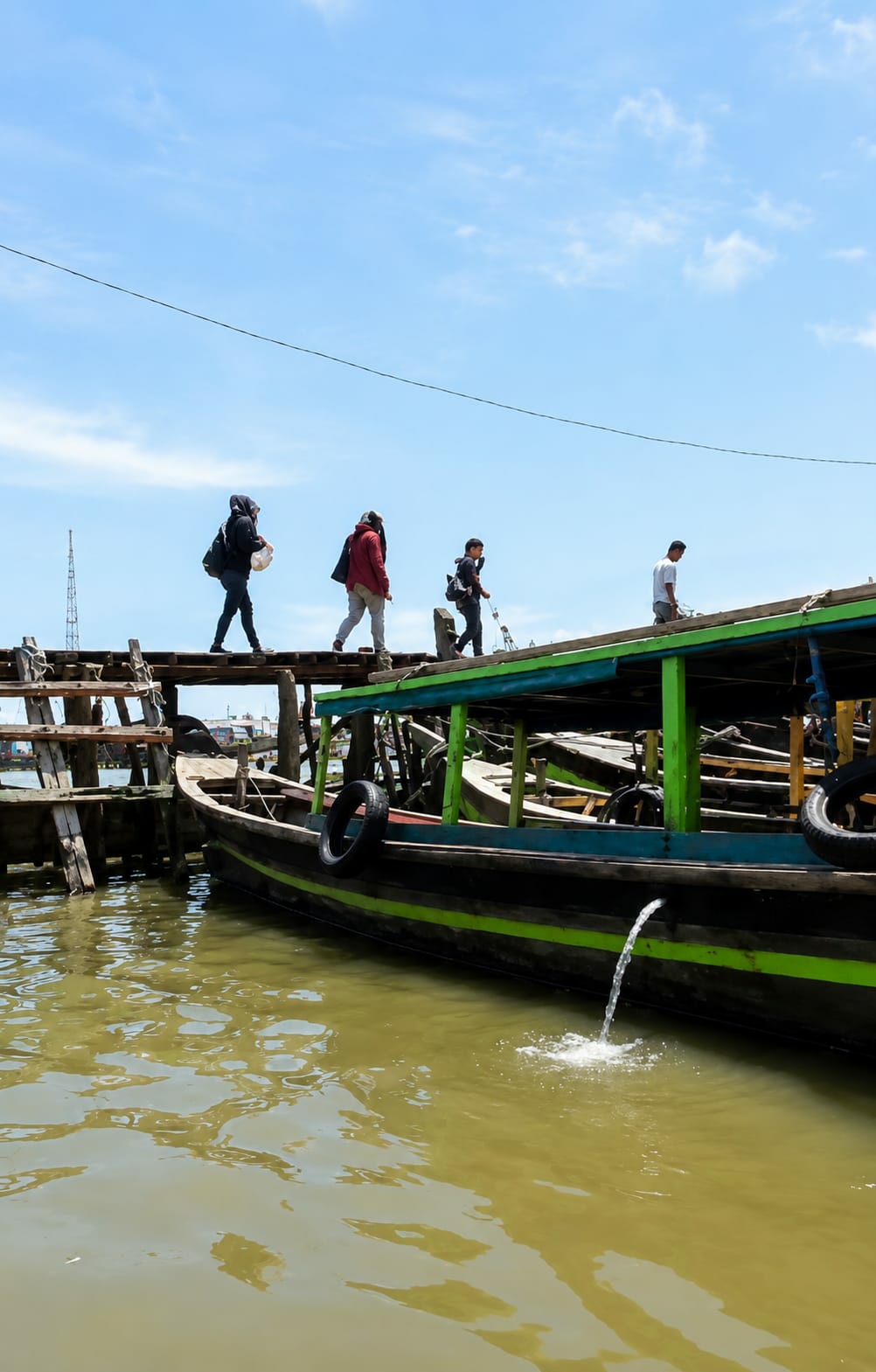 In the City of Medan, Indonesia, on a bright, partly cloudy day, a wooden pier extends over murky green water. Four individuals are walking across the pier. From left to right, the first person is wearing a dark hooded jacket and carrying a white bag in their left hand and a dark backpack over their right shoulder. The second person wears a red jacket and dark pants, carrying a white object in their right hand and a dark backpack over their left shoulder. The third person, a youth, is dressed in dark clothing with a dark backpack. The fourth person, a man, wears a white t-shirt and dark pants. To the right of the pier, a long, dark green and lime green boat is docked, its wooden structure visible, along with two black tires mounted on its side. Water is seen gushing from an opening on the boat's hull into the water below. In the background, several other boats are visible, some partially obscured by debris. A tall, thin radio tower stands to the far left of the frame.