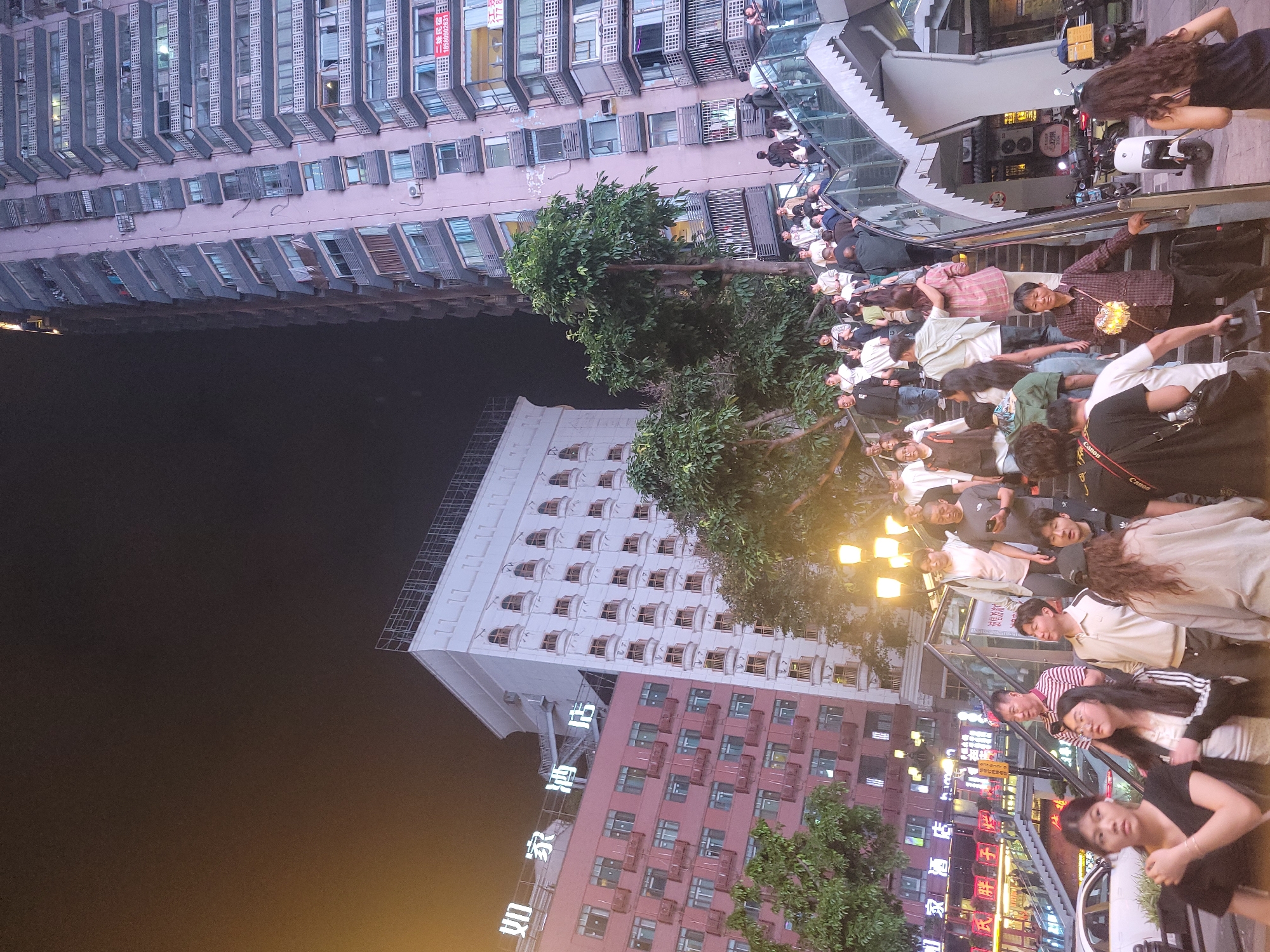 This image captures a vibrant night scene in an urban area of Yuzhong District, China, featuring multi-story buildings, a bustling crowd, and illuminated signs. The perspective is slightly tilted, looking up at the high-rises.
In the background, several tall buildings dominate the skyline under a dark night sky. To the left, a light purple/pink high-rise building with numerous windows, many featuring external grates or shutters, stretches upwards. A red sign with white Chinese characters and a partially visible phone number "0509092631" is affixed to this building, likely advertising property for sale or rent. Below this, a white building with distinctive arched window frames is visible, and further to the right, a darker red/brown building with modern windows. Luminous white Chinese characters reading "如家酒店" (Home Inn Hotel) are vertically displayed on the side of the white building.
The foreground is filled with a large crowd of people, mostly young and middle-aged adults, gathered around and on a set of escalators or stairs with glass railings. People are either ascending, descending, or standing, creating a dynamic sense of movement and activity. Their attire is casual. One person in the middle ground holds a sparkling, glowing object. Other individuals are looking towards the camera. Near the top right, a white scooter or motorcycle is parked. Trees with green foliage are interspersed within the urban environment, and streetlights illuminate parts of the scene with warm light. Various other neon and illuminated signs with Chinese characters are visible on the lower buildings, contributing to the lively night ambiance. A "Canon" logo is visible on a camera strap or bag worn by one person. The overall impression is one of a busy, modern city at night.