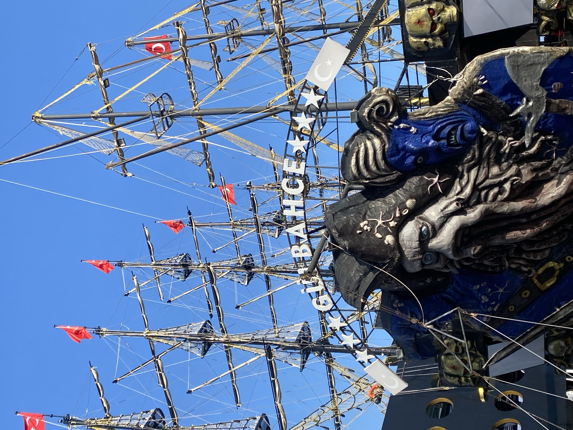 This image showcases a detail-rich view of a pirate-themed ship or a maritime attraction against a bright, clear blue sky. The vessel features multiple dark masts with extensive rigging and a network of ropes and nets, designed to evoke the appearance of an old sailing ship.
In the foreground, a large and intricately sculpted decorative element, possibly a figurehead or a section of the stern, dominates the right side of the frame. It depicts a formidable sea creature or monster, characterized by a grey, textured face with piercing blue eyes, tentacles, and other marine-inspired details, painted in shades of blue, grey, and black. Above this, other decorative elements resembling golden skulls with red eyes are visible.
Several flags are present: small red pennant flags flutter from various points on the rigging, adding splashes of color. Additionally, white flag-like banners, each displaying the red crescent moon and star emblem of the Turkish flag, are attached to the rigging and a vertical sign. This vertical sign features a line of white stars and partially visible text that reads "GIZLIB BAHCE."
The scene is illuminated by bright daylight, under a cloudless, vivid blue sky, indicating fair weather. The overall aesthetic suggests an adventurous and dramatic theme, consistent with pirate lore. Based on the visible Turkish flags and the provided context, the location is Alanya, Turkey, likely a popular tourist attraction or tour boat. No people are visible in the image.