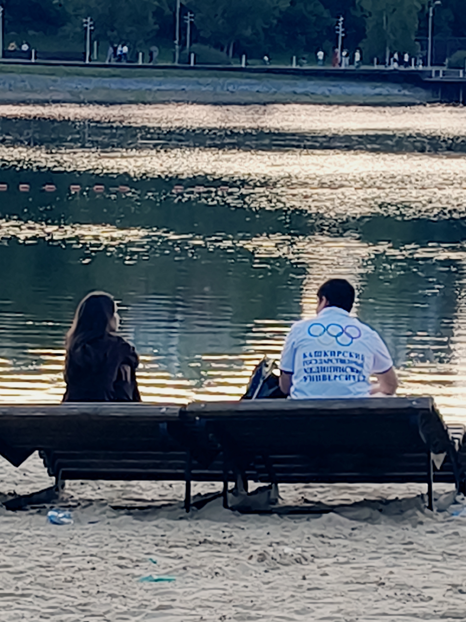 Two individuals, a woman and a man, are seated on a public bench facing a body of water. The woman, with long dark hair, is on the left, looking to her right. The man, on the right, is wearing a white polo shirt with the Olympic rings emblem and Russian text, and is looking forward. The bench is situated on a sandy beach, with trees and a sidewalk visible in the background across the water. The water is calm with reflections of sunlight.