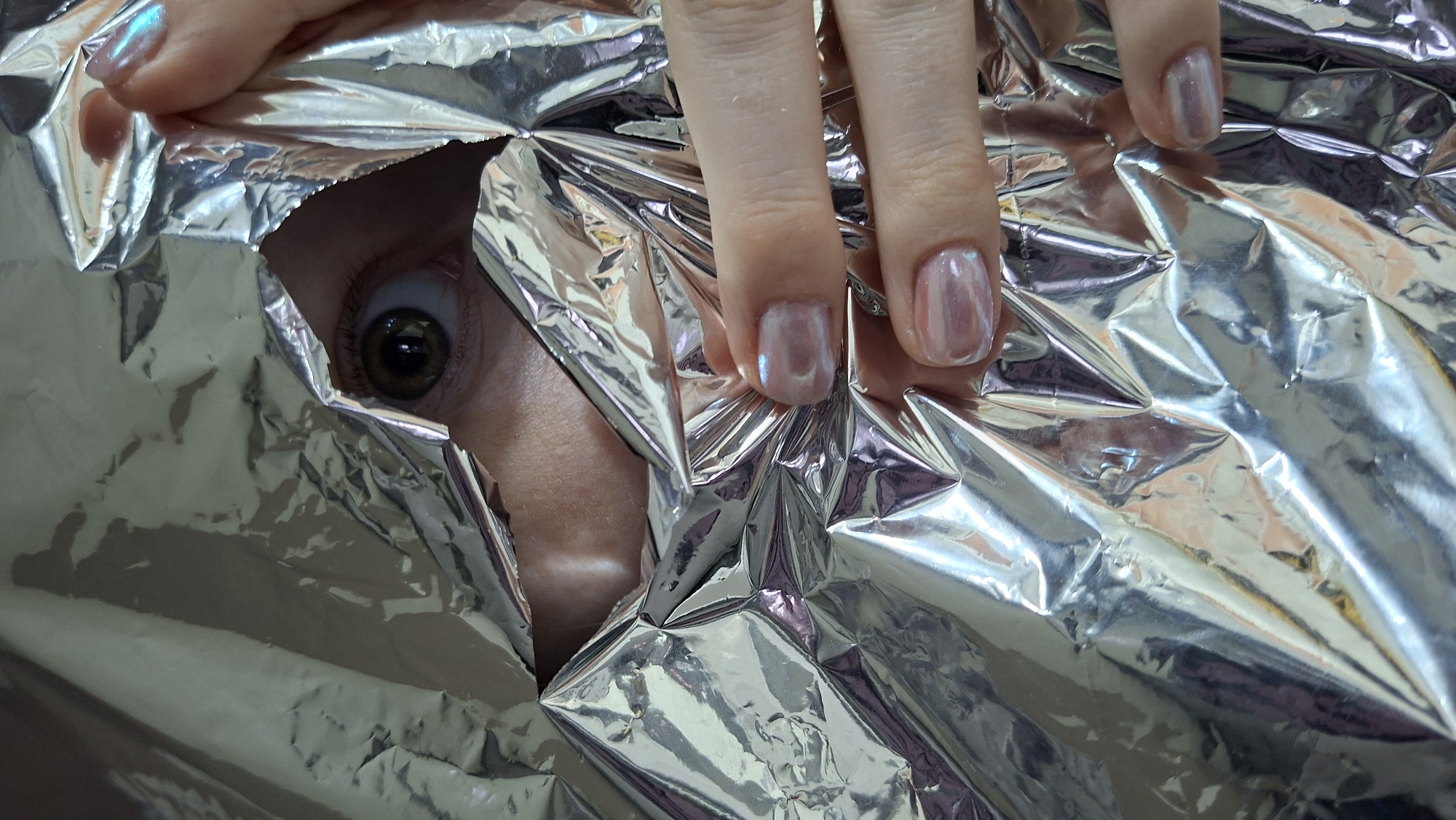 A person is seen peering through a hole in a sheet of shiny, reflective material (possibly aluminum foil). Only one eye is visible, along with part of the face around the eye. A hand with painted fingernails is positioned near the opening, seemingly holding the foil in place. The foil is crumpled and reflects light, creating a textured and distorted appearance. The location mentioned is Chaikovsky, Russia, but there's no visual information within the image itself to confirm the location.