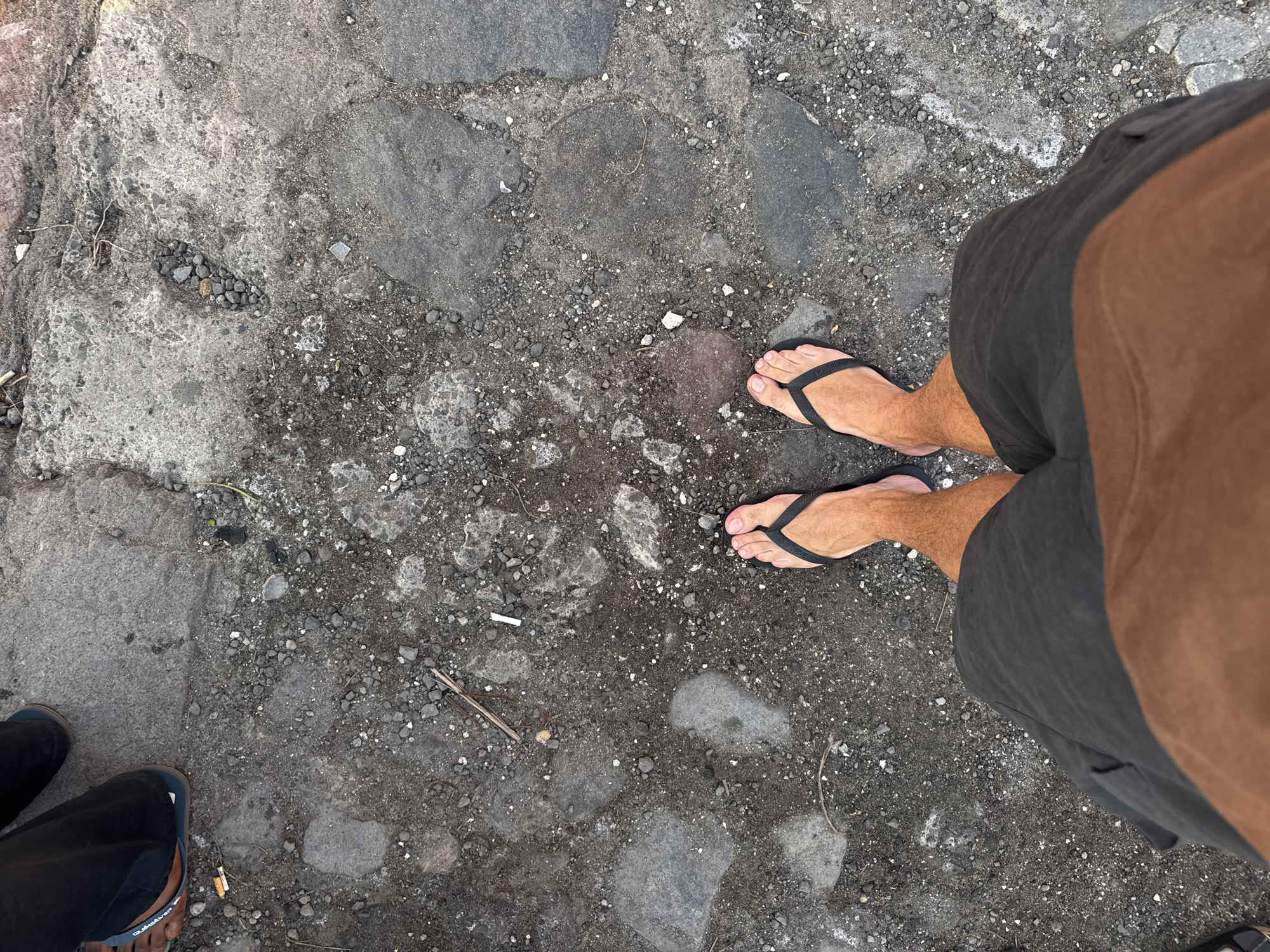 The image shows a view from above of a person standing on a stone paved surface. The person is wearing a pair of black flip-flops and grey shorts. The stone pavement consists of irregular-shaped stones of varying sizes and shades of grey. The pavement is also covered with small stones, dirt, and bits of organic matter. Another person's legs and feet are visible in the bottom left corner of the image, wearing blue sandals. The location is North Kuta, Indonesia.