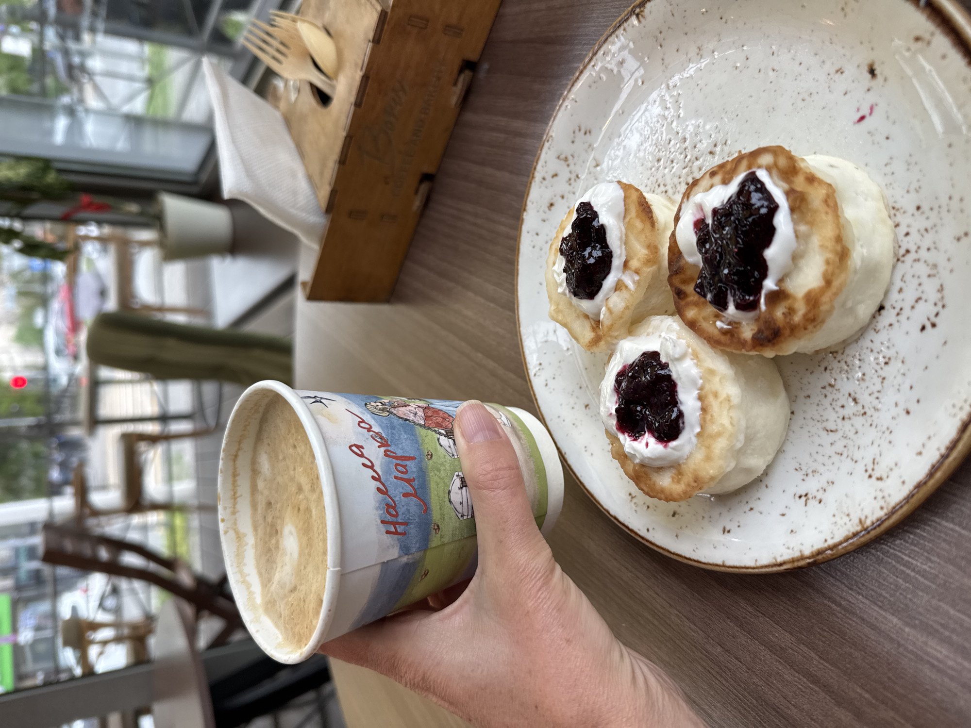The image captures a first-person perspective of a cafe setting, likely in Bishkek City, Kyrgyzstan, focused on a breakfast or snack. In the immediate foreground, a hand holds a paper coffee cup filled with a light-brown beverage, likely a latte, featuring a faint foam design. The cup is adorned with colorful illustrations of a rural landscape and handwritten-style Cyrillic text that reads "Наслаждайся Мармелад" (Enjoy Marmalade/Jam).
To the right, a rustic-looking ceramic plate with speckled patterns holds three round, pan-fried pastries, resembling traditional Syrniki (cottage cheese pancakes). Each is generously topped with a dollop of white cream or sour cream and a dark, glossy berry jam or compote.
In the upper-left, a wooden caddy, designed to hold condiments or utensils, contains several wooden forks and spoons, along with white paper napkins. Faint, burned-in text on the side of the caddy appears to read "COFFEE AND BAKERY." The items rest on a light brown wooden tabletop.
The background is blurred, showing large windows through which an urban outdoor scene with some greenery is visible, indicating a daytime setting. The overall impression is one of a relaxed meal in a brightly lit cafe.