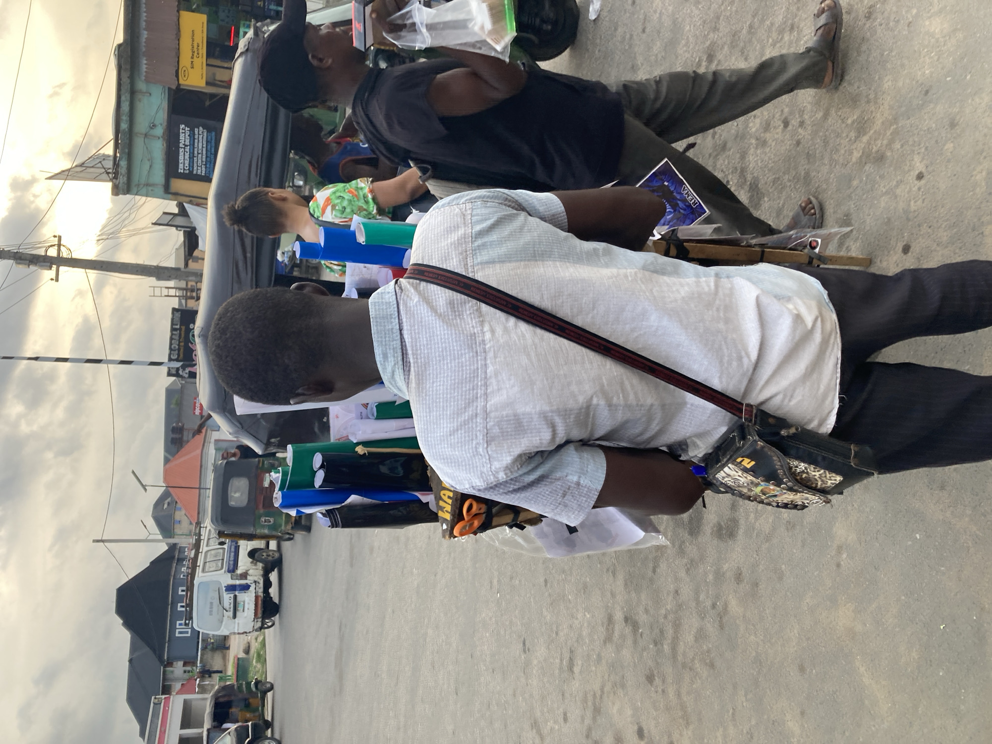 The image shows a street scene in Aba, Nigeria. The foreground features a person with a striped white shirt and a bag with a leopard-print design slung over their shoulder. The person is carrying rolls of colorful vinyl material. Behind this person, another individual in a black shirt and cap is standing, holding a plastic bag. In the background, there are auto rickshaws parked along the street and several buildings with signs like "ZIKSONS PAINTS & CHEMICAL DEPOT", "NTN SIM Registration Center" and "GLOBAL LINK". The sky is overcast. The scene is captured at street level, showing the urban environment of Aba.