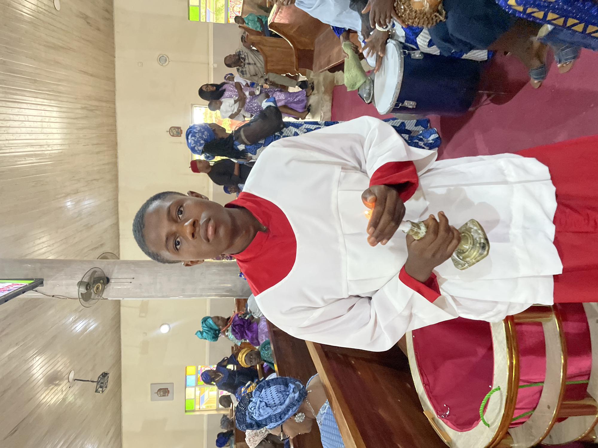 The image depicts the interior of a church in Gura Top, Nigeria. A young man in a white and red robe, possibly an altar server or religious figure, is holding a small candlestick or chalice. He is positioned in the foreground, looking directly at the camera. The church pews are filled with congregants, mostly women, some wearing colorful headwraps. A drum is visible in the foreground, indicating music as part of the service. The walls are light-colored and feature horizontal paneling. There are stained glass windows, ceiling fans, and a flat-screen television mounted on the wall. The overall atmosphere is one of a religious service in progress.