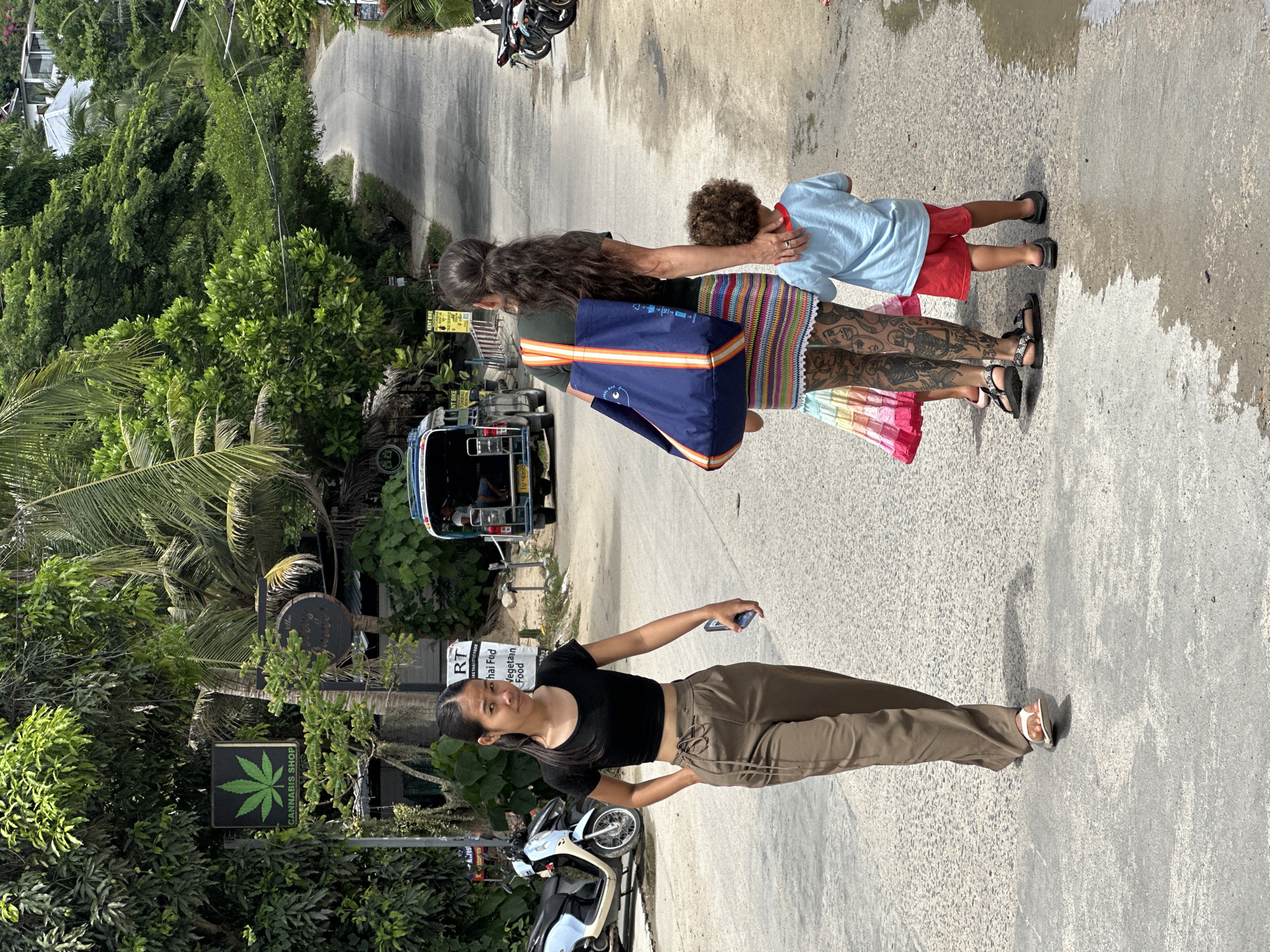The image depicts a street scene in a tropical location, likely ตำบลเกาะพะงัน, Thailand, characterized by lush greenery and a paved road. In the foreground, a young woman with dark hair pulled back in a ponytail is walking towards the viewer, holding a smartphone in her right hand. She is wearing a black crop top and loose-fitting, brown, high-waisted trousers. To her left, a light blue and white scooter is parked on the side of the road.
Further down the road, a woman with long, dark, wavy hair is walking away from the camera, holding a large blue tote bag with orange and white stripes over her right shoulder. She is wearing a dark green t-shirt and a brightly colored, striped skirt. A young child with curly brown hair, wearing a light blue t-shirt and red shorts, walks beside her, with the woman's hand resting on the child's shoulder. Another child, partially visible, is wearing a pink and yellow dress.
In the background, a turquoise and silver tuk-tuk is parked on the left side of the road. Several motorcycles are parked on the right side of the road further up. Overhead, power lines are visible, and the road curves to the right, leading into a more densely vegetated area. A sign with a green cannabis leaf and the words "CANNABIS SHOP" is prominently displayed on the left side of the road. Another sign with "RT THAI Food VEGETARIAN FOOD" is also visible. The ground is a mix of paved road and some damp patches, suggesting recent rain or water runoff. The overall atmosphere is sunny and warm, typical of a tropical climate.