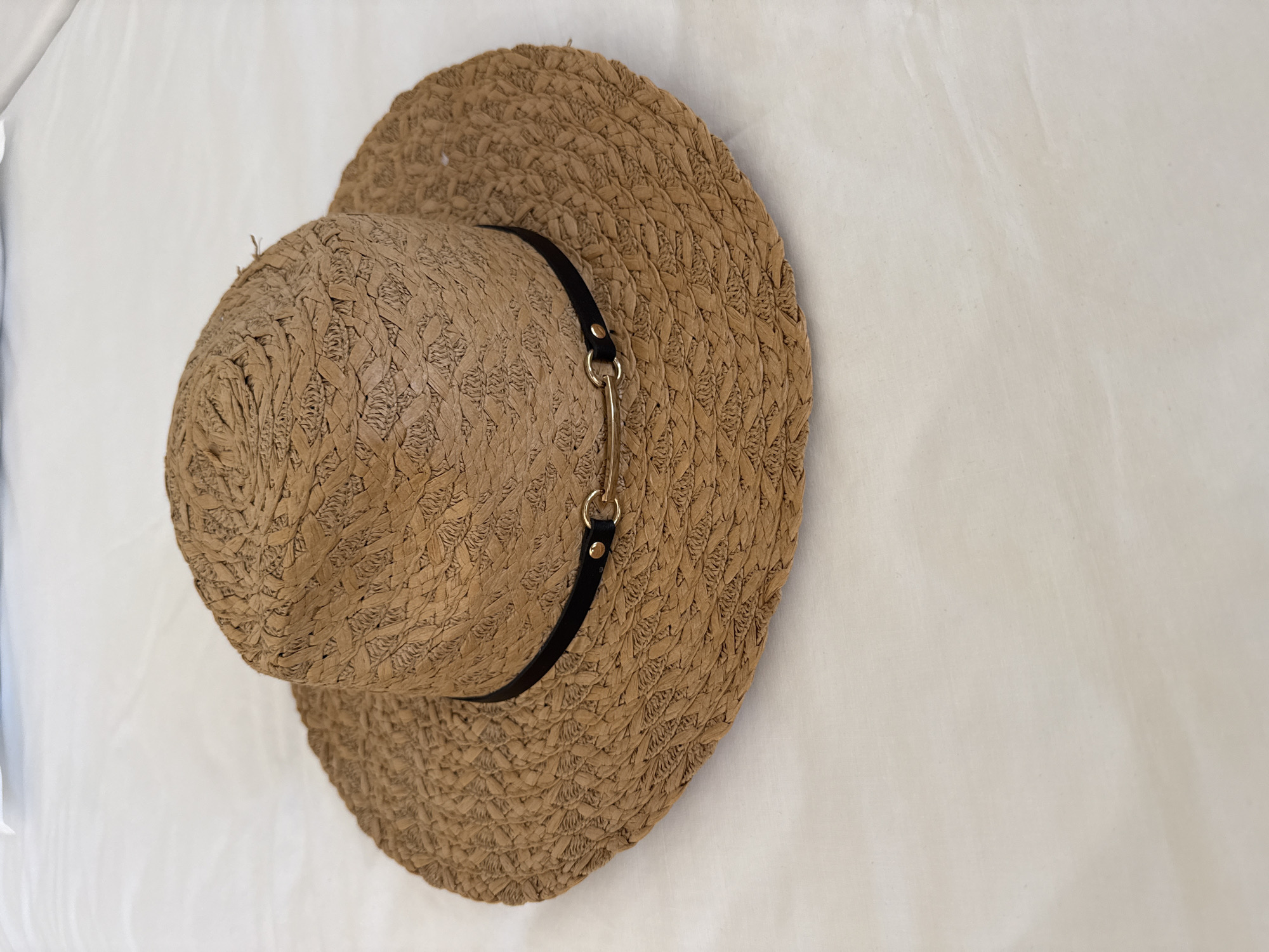 A woven straw hat with a black band and gold buckle sits on a white, rumpled surface. The hat is light brown with a patterned weave. The surface appears to be fabric, possibly a bedspread or tablecloth, with soft folds and shadows. The lighting is even, suggesting it was taken indoors. There are no people or visible actions occurring. The hat is the sole subject of the image. The location is not specified in the image or its metadata.