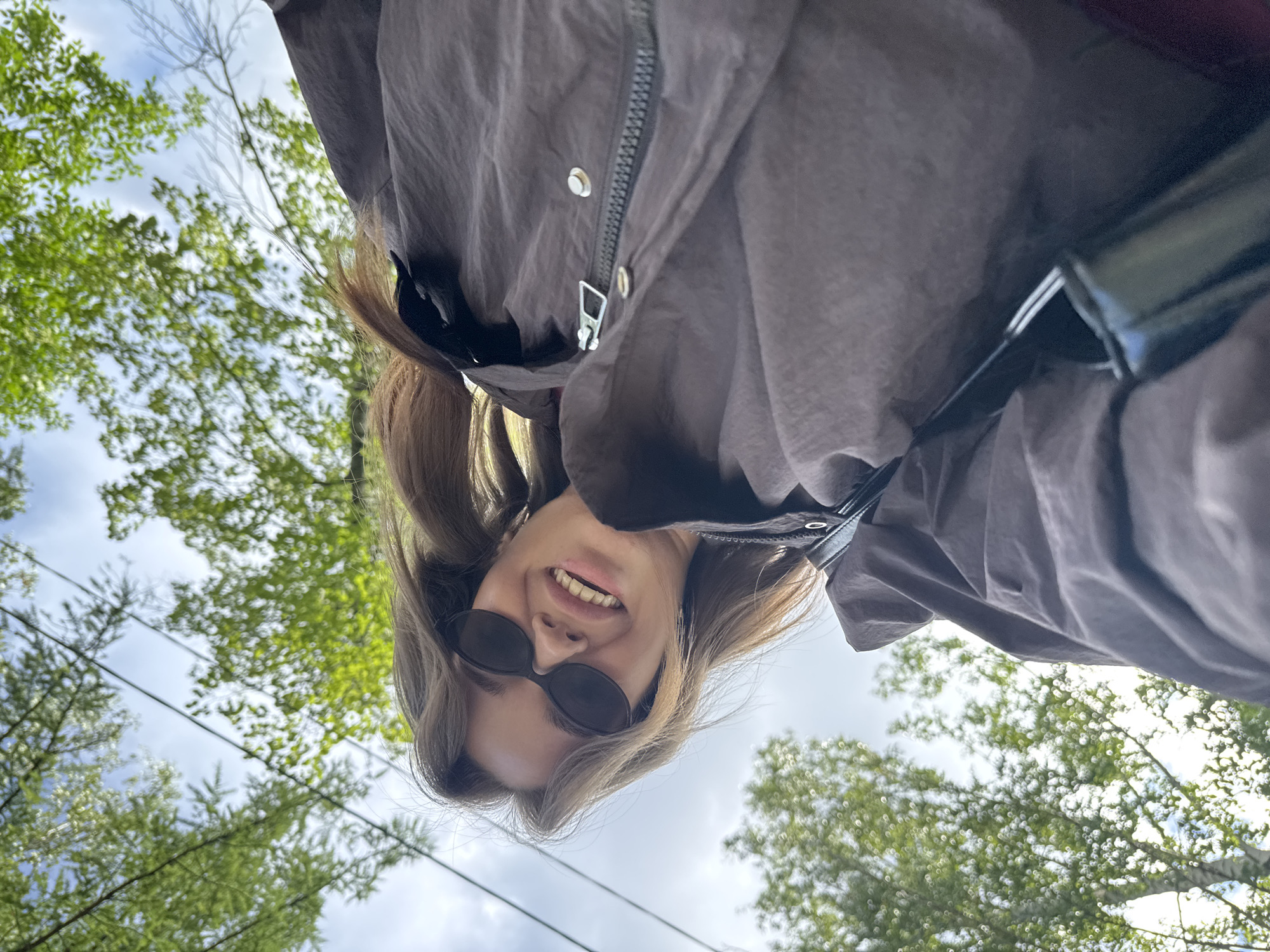 The image, taken outdoors in Chaikovsky, Russia, is an upside-down selfie of a young woman. She is wearing dark sunglasses and a brown jacket with a zipper and metal snap buttons. Her hair, which is a light brown color, is falling around her face. She is smiling, with her teeth showing.
The background is a mixture of green trees and a cloudy sky. The perspective suggests the photo was taken from a low angle, looking upwards towards the woman and the sky. The presence of trees and the cloudy sky suggest it might be daytime, possibly overcast. There are some thin, dark lines visible in the sky, which could be wires or branches. The overall mood is casual and lighthearted, indicated by the woman's smile and the outdoor setting. No visible text is present in the image.