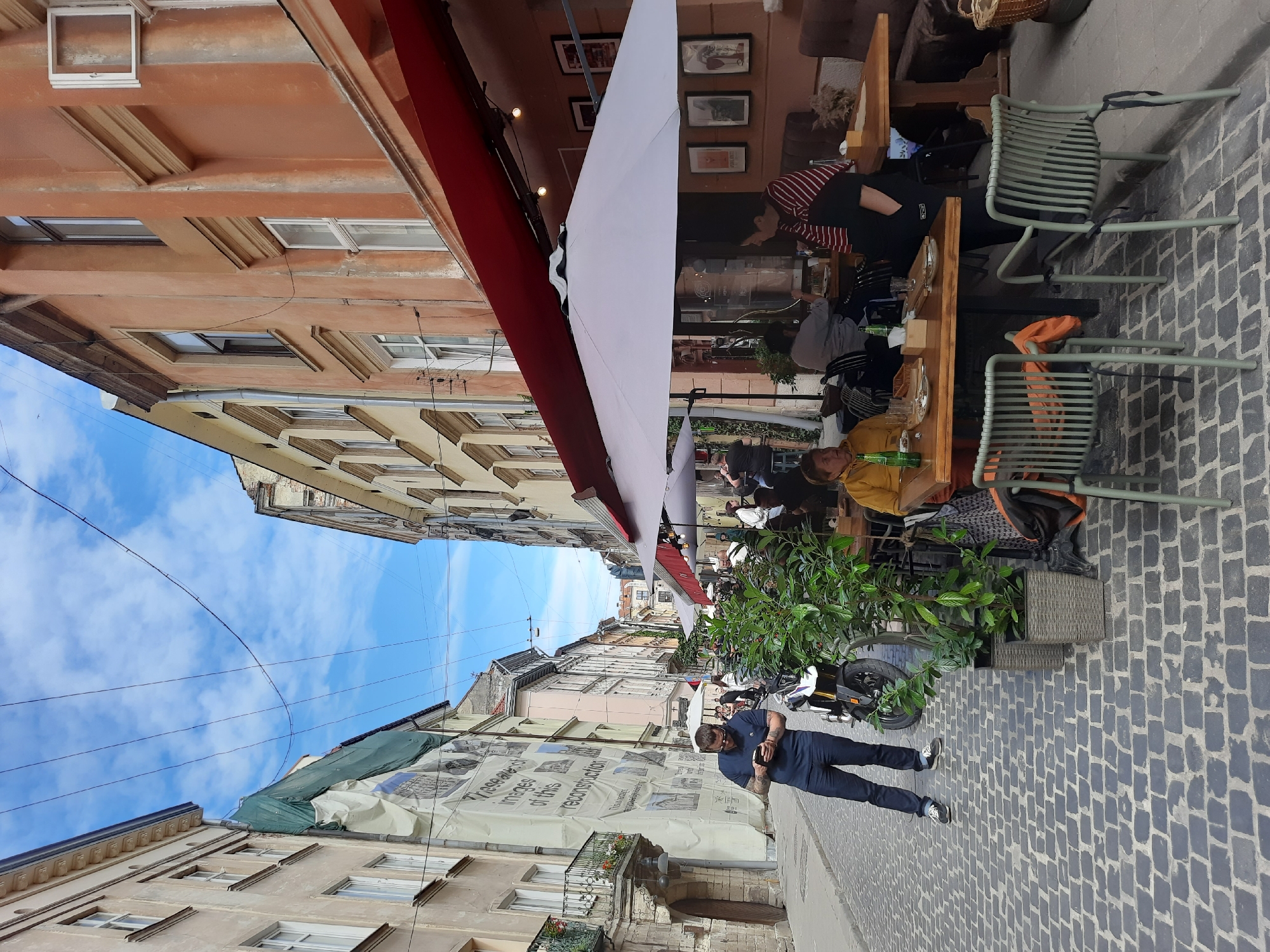 A sunny daytime exterior shot captures a cobblestone street in Lviv, Ukraine, lined with multi-story buildings. The street is partially occupied by an outdoor seating area of a cafe, complete with umbrellas and tables. A man stands on the left, looking at his phone, near a motorcycle and some potted plants. Several patrons are seated at the cafe tables, either interacting or looking away from the camera. The buildings are a mix of architectural styles, with some appearing older and others more recently renovated. The sky is blue with scattered clouds.