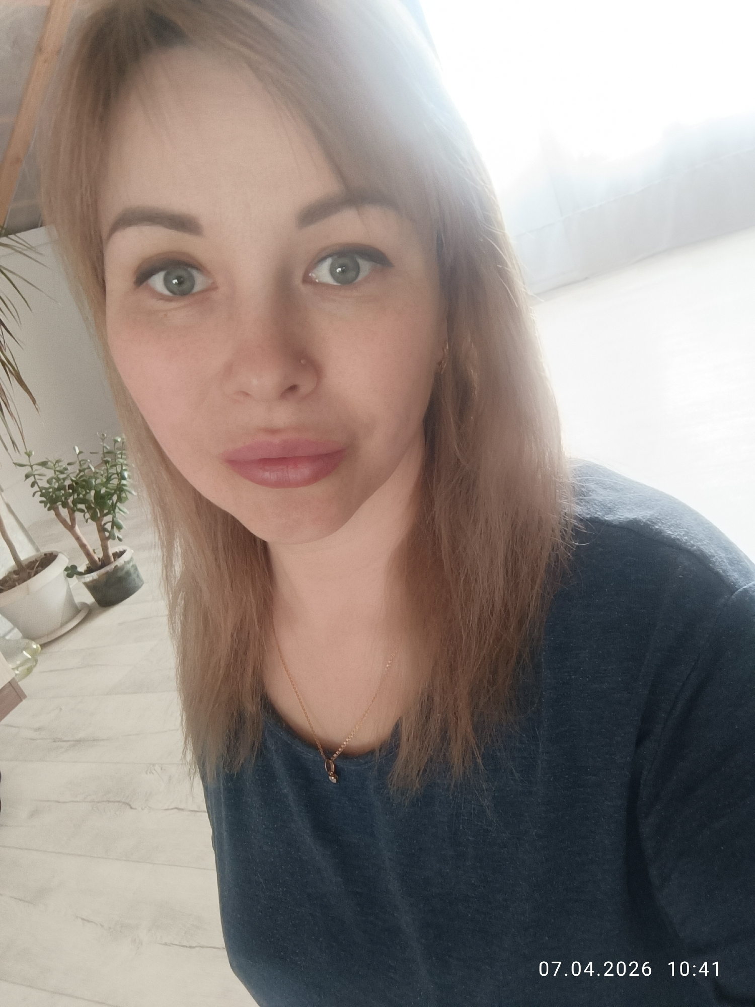 A medium-angle, bright, close-up selfie of a woman. She has blond hair and green eyes. She is wearing a dark blue t-shirt and a gold necklace. In the background are two plants in pots, a white wall, and a pale wooden floor. The image includes a timestamp of 07.04.2026 10:41. The city of Chaikovsky, Russia, is not identifiable from the image.