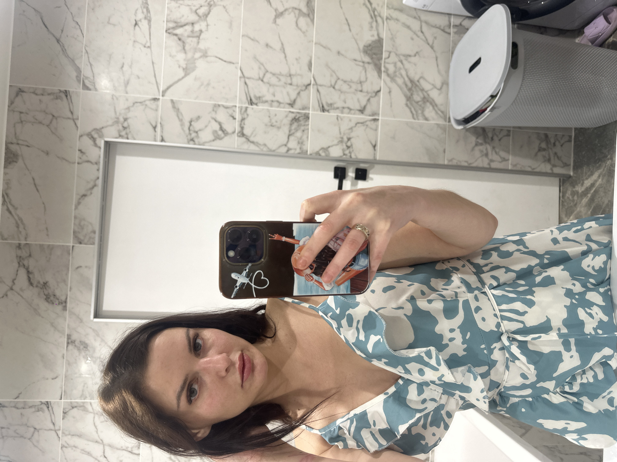 A woman with long, dark hair is taking a mirror selfie in a bathroom. She is wearing a blue and white patterned dress with ruffles. Her left hand is holding her phone, which has a patterned case with a picture of a person on a beach. Her right hand is holding her hair. The bathroom has marble-tiled walls and a white door. A laundry hamper is visible to the right of the woman.