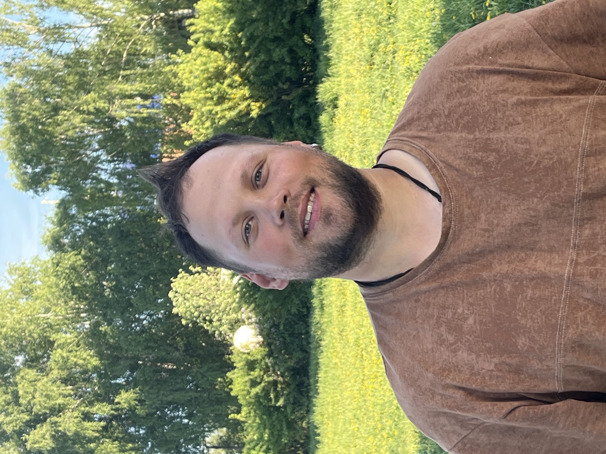 A man is standing outdoors in a grassy area with trees in the background. The man has a beard and short hair, wearing a brown shirt. The trees and grass are green, and the sky is visible in the upper left corner. The image is taken at an angle. There is no evidence to suggest this photo was taken in Фоки, Russia.