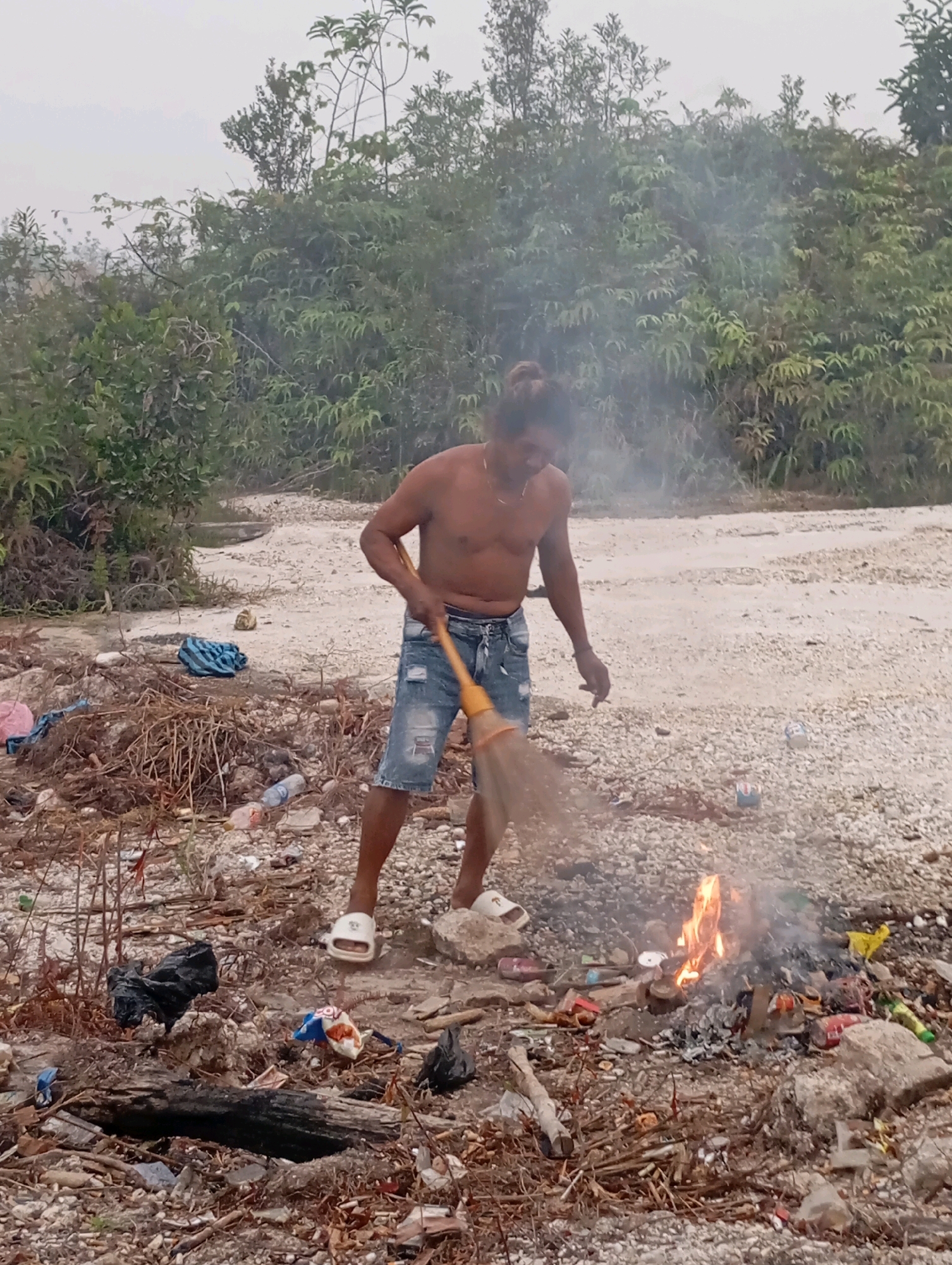 An adult male, shirtless and wearing denim shorts and white slides, is depicted in a semi-outdoor environment with dense vegetation in the background and a gravelly, debris-strewn ground in the foreground. The man is holding a broom with an orange handle and appears to be sweeping near a small fire. The fire is actively burning, producing smoke, and is surrounded by various items of refuse, including plastic bottles, cans, and wrappers. The ground is covered with small stones, dry branches, and a significant amount of litter. In the background, a dirt road or path is visible, bordered by the vegetation. The overall impression is a scene of outdoor cleanup or waste disposal in a somewhat unkempt area.