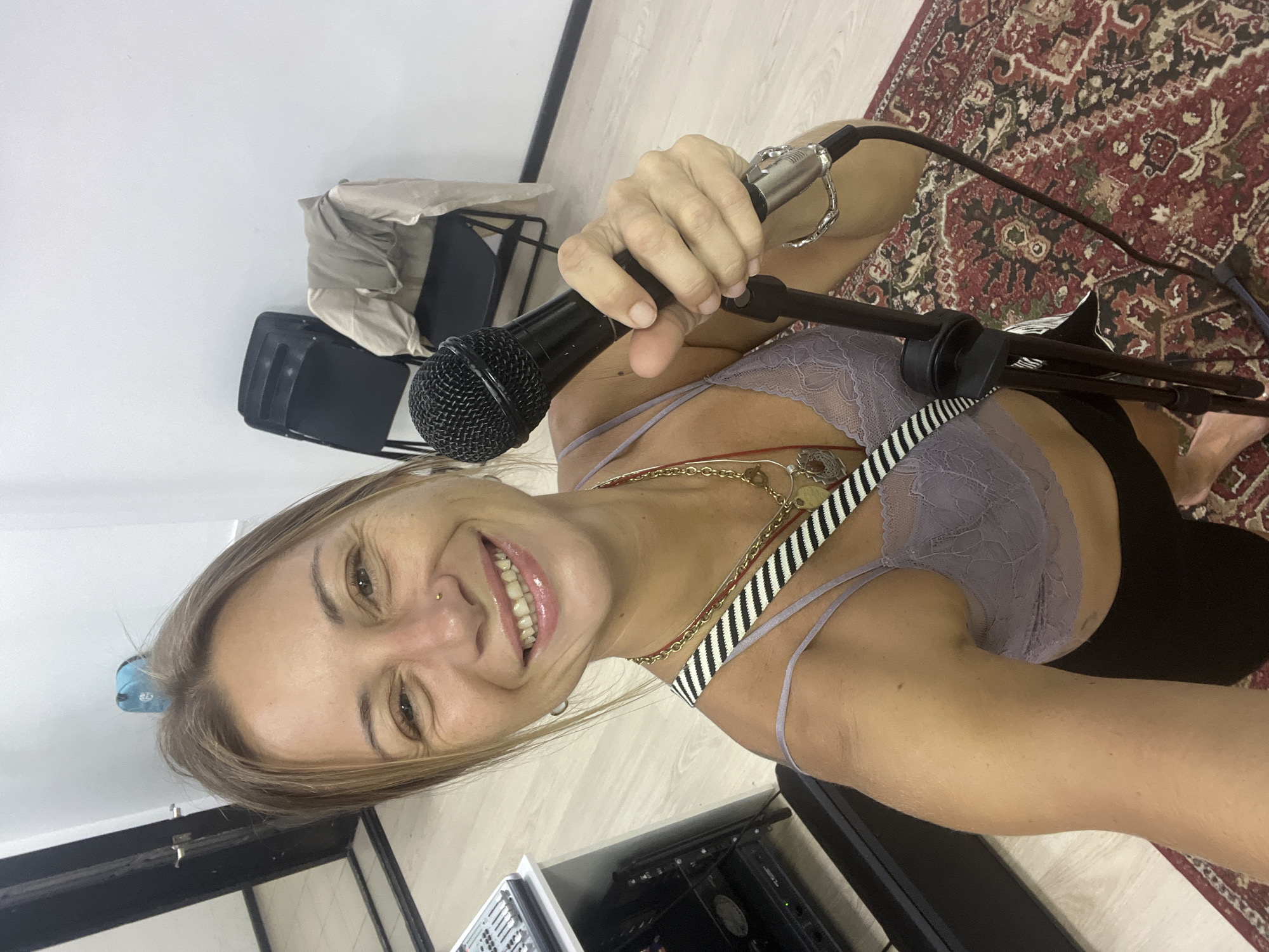 A young woman with blonde hair is holding a microphone in a room that appears to be a studio or rehearsal space. She is smiling and looking at the camera. She is wearing a lacy purple bra and several necklaces. In the background, there are two black chairs, one of which is covered with a light-colored cloth, and a colorful patterned rug on the floor. There is also some audio equipment visible. The lighting suggests it could be daytime indoors. The overall mood is cheerful and energetic. There is no visible text in the image.