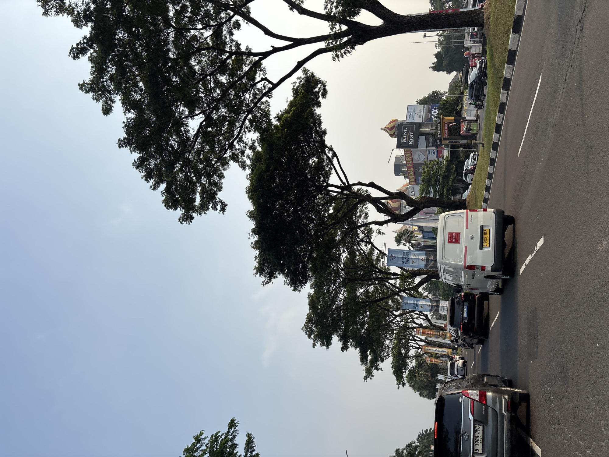 The image shows a street scene in Pakulonan Barat, Indonesia, likely during the daytime with a clear sky. A line of vehicles is on the road, with a silver SUV in the foreground on the left, followed by a white van, and other cars further down the road. Trees line the side of the road, casting shadows. In the background, there are several buildings with signs, including "HANDAL FURNIT", "KING KOIL", and "THERAPEDIC". Banners with "ONE DISTRICT TWO ICONS" are also visible. The road has clear lane markings.