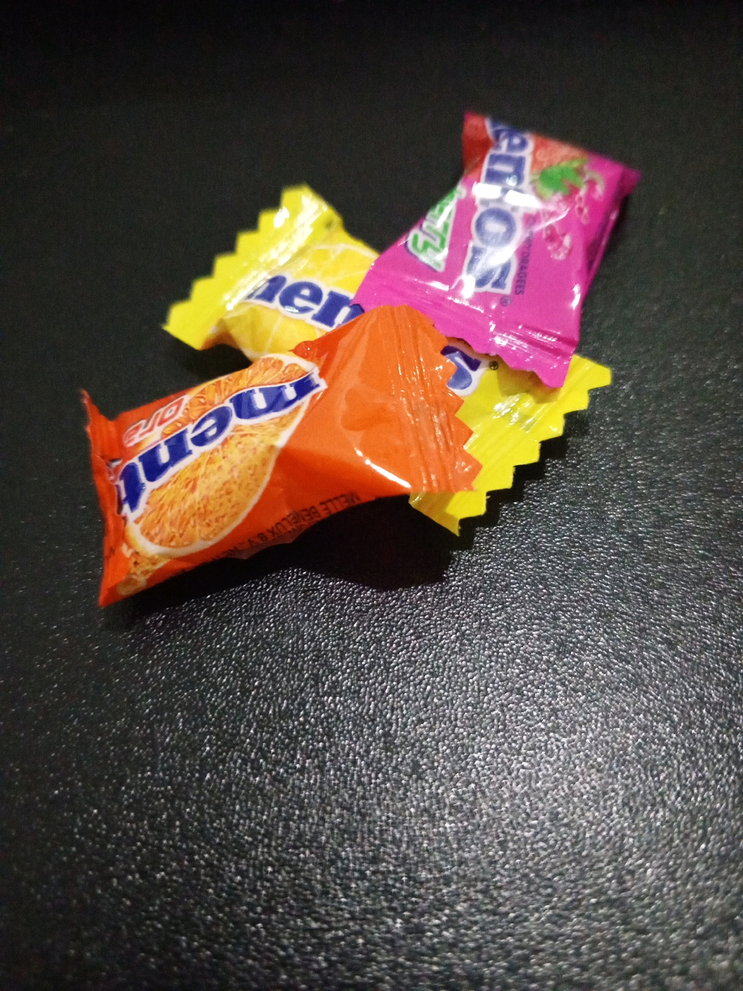 Three individually wrapped candies are arranged on a textured dark surface. The candy on the bottom left is orange and features a depiction of an orange slice with the word "Mentos" in blue lettering. To its left and slightly behind is a yellow-wrapped candy, also with "Mentos" visible. Overlapping the other two, and positioned towards the top right, is a pink-wrapped candy with "Mentos" printed in white and blue, accompanied by an image of red berries. The lighting suggests an indoor setting, with reflections visible on the candy wrappers. The surface appears to be a table or countertop. There are no people visible in the image, nor any indication of the city of Denpasar, Indonesia.