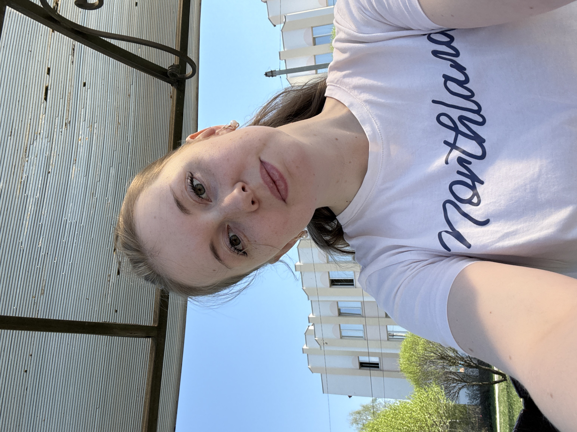 A young woman is pictured from a slightly low angle, looking at the camera. She has fair skin, light brown hair pulled back, and is wearing a white t-shirt with the word "Northland" written vertically in navy blue script. Behind her is a combination of outdoor and architectural elements. To her left, a corrugated metal structure with a dark metal frame is visible. In the background, a light-colored building stands against a clear blue sky. Below the building are trees with green foliage. The location is the city of Unknown, Unknown.