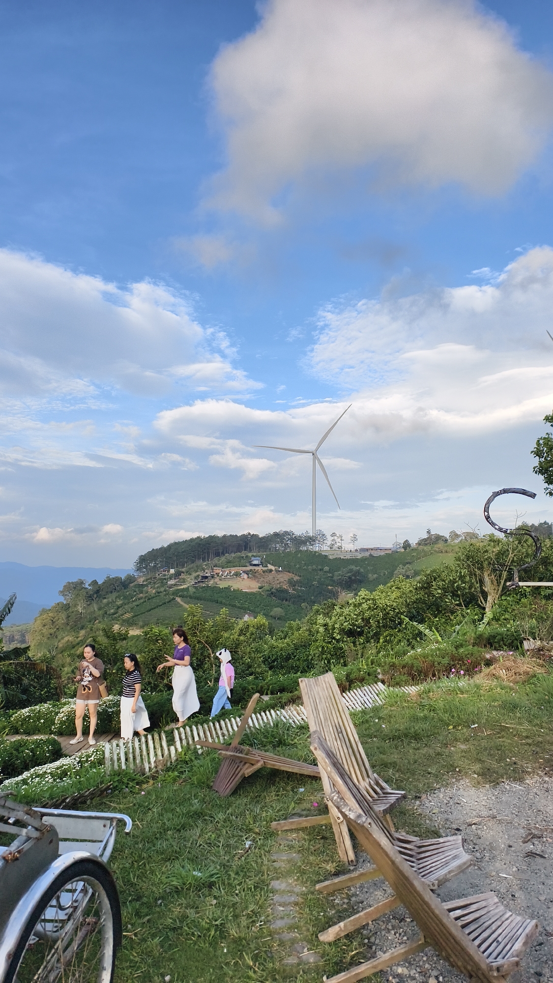 This image shows a scenic landscape in Phát Chi, Vietnam. In the foreground, two wooden chairs are laid on the ground next to a pathway. Further back, a group of four people are walking along a white wooden fence that separates a garden area with white flowers from a grassy slope. The people appear to be on a tour or enjoying the view. Behind them, the landscape unfolds with rolling green hills covered in trees and vegetation, indicative of tea or coffee plantations. A prominent wind turbine stands tall on one of the hills, suggesting a commitment to renewable energy in the area. The sky is partly cloudy, with dramatic white clouds against a blue expanse. The overall scene captures a serene and picturesque rural environment.