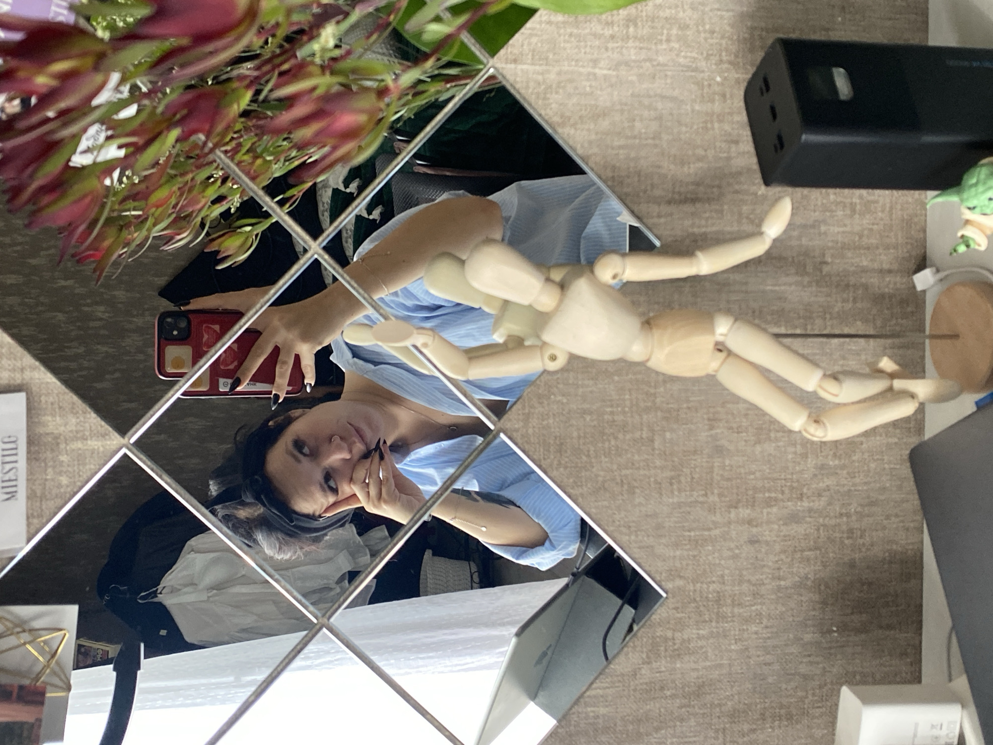 A woman is taking a mirror selfie in a room. She is wearing a light blue striped shirt and a black headband. She is leaning her head on her hand and looking at her phone, which she is holding in her other hand. In the mirror, her reflection is visible, along with the room behind her. The room appears to be a home office or living space, with a desk, a laptop, and a vase of flowers. A wooden mannequin is also visible in the foreground, in front of the mirror. The image is taken from a medium shot, focusing on the woman and her reflection.