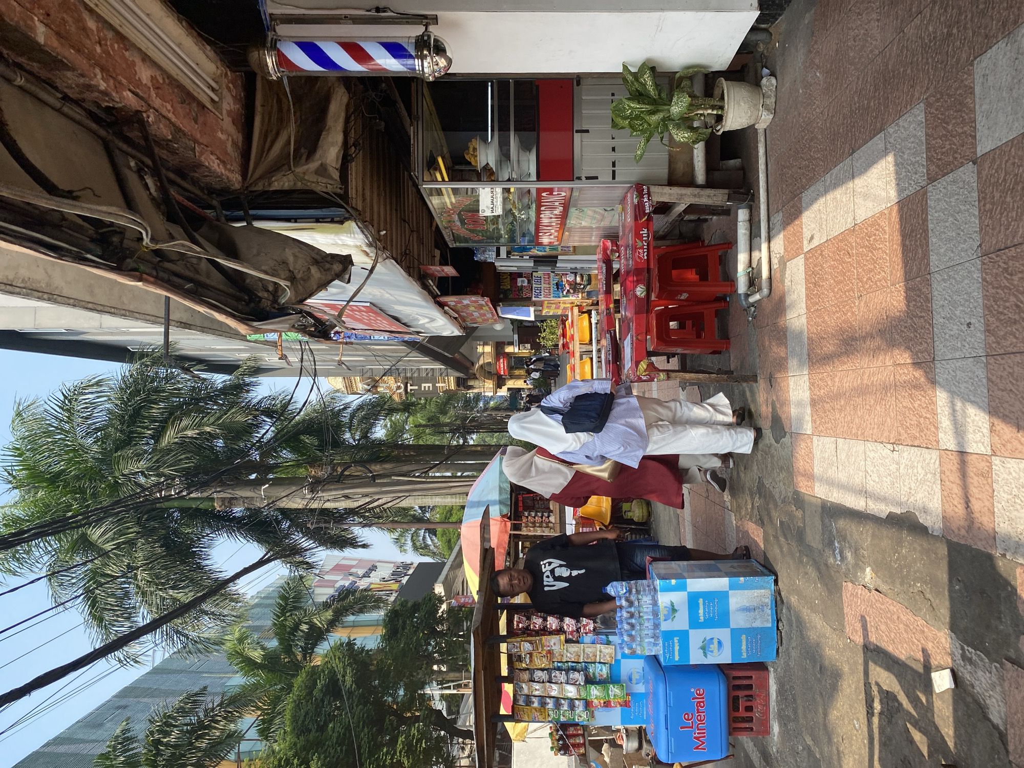 The image depicts a street scene in South Jakarta, Indonesia, likely near a commercial area. In the foreground, a sidewalk with a checkered pattern of light brown and beige tiles runs alongside a row of shops. To the left, a street vendor's stall is set up, displaying various packaged goods, bottles of water, and two blue coolers. A man wearing a black t-shirt stands in front of the stall, facing the camera.
Further down the sidewalk, three people are walking away from the camera. Two are wearing hijabs, one in a burgundy garment and the other in white. The third person is wearing white pants and a light blue and white striped top.
Adjacent to the vendor's stall is an outdoor seating area with red tables and chairs, set up for dining. Signs for "MASAKAN PADANG" (Padang cuisine) are visible on the storefront, indicating a restaurant. A barber pole with red, white, and blue stripes stands outside another establishment.
The environment is characterized by tall palm trees lining the street, utility poles with numerous electrical wires, and modern buildings in the background, one of which has a glass facade. The lighting suggests it is daytime, with shadows cast by the trees and structures on the sidewalk.