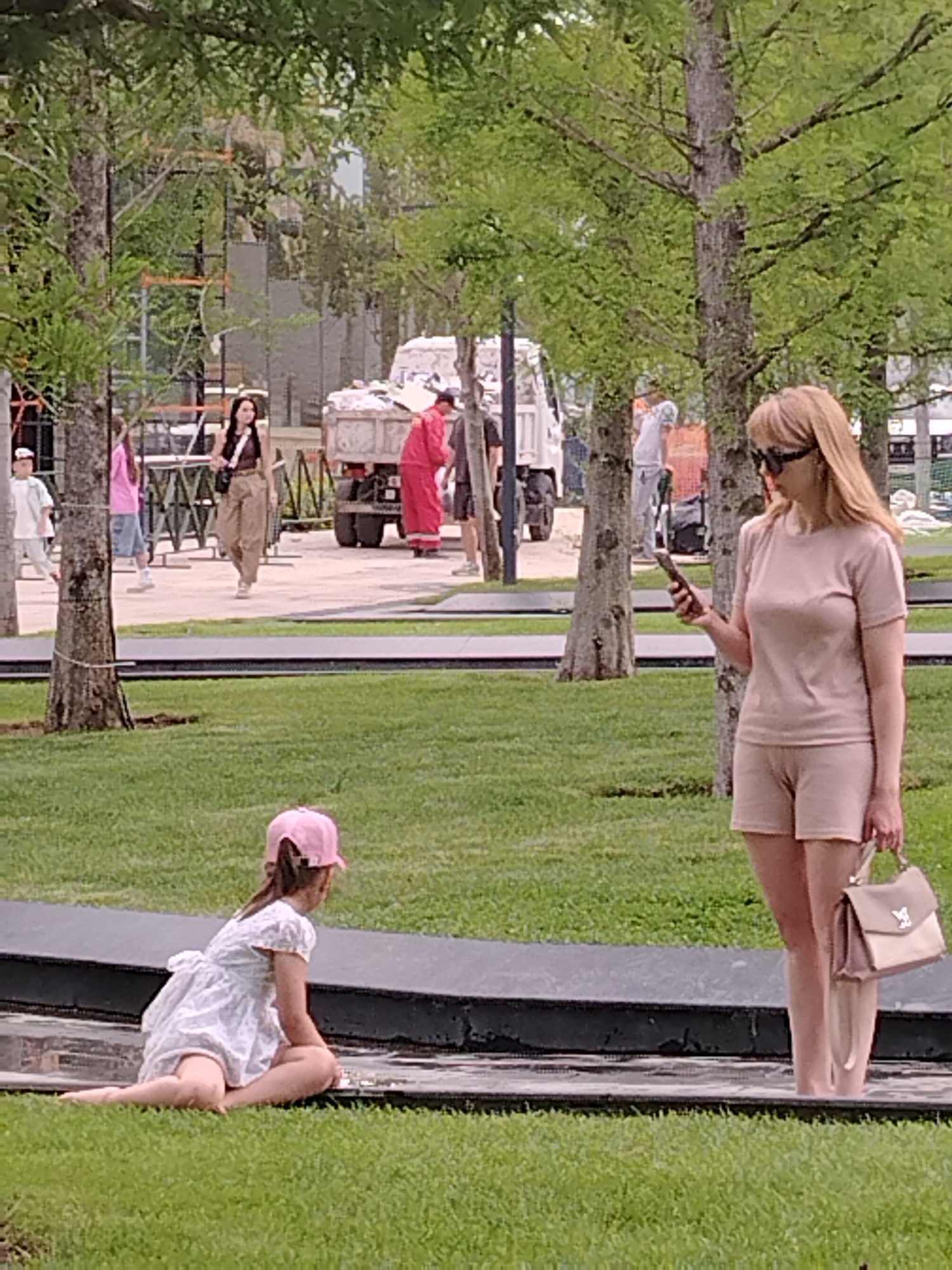 In Krasnodar, Russia, on a cloudy day, a woman with blonde hair and sunglasses stands in a park, holding a smartphone in her right hand and a handbag in her left. She is wearing a light pink t-shirt and shorts. In the foreground, a young girl wearing a white dress and a pink baseball cap sits on the edge of a dark, water-filled planter, facing away from the camera. In the background, several trees line a grassy area, and a truck and other people are visible.