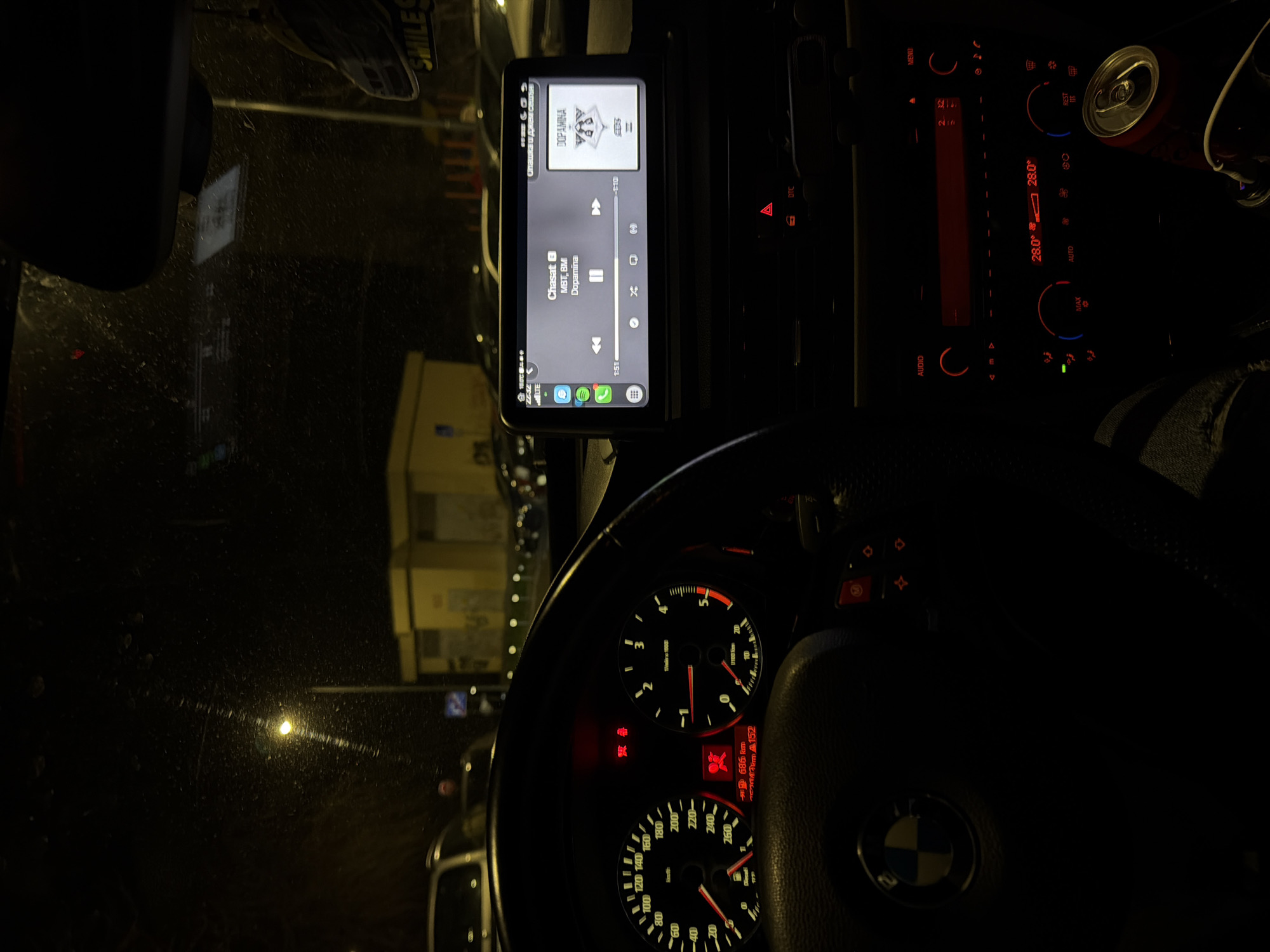 The image depicts the interior of a BMW vehicle at night, viewed from the driver's perspective. The illuminated instrument cluster shows a speedometer indicating 0 km/h and a tachometer at 0 RPM, with a digital readout displaying 686 km. Multiple red warning lights are active, including those for an unbuckled seatbelt and an airbag malfunction. An aftermarket infotainment screen is mounted above the factory radio and climate controls. The screen displays a Spotify interface playing "Chasat" by "MBT, BMI" with the album title "Dopamina," showing a timestamp of 1:51 into the track and a total duration of 4:01. The screen also indicates "LTE" network connectivity and the time "22:42". Below the screen, the red-backlit climate controls show a temperature setting of 28.0°C. In the lower right corner, a red beverage can and a white charging cable connected to a car charger are visible. An air freshener, shaped like a car with "SMILE" text, hangs from the rearview mirror.
Through the visibly dirty or rain-speckled windshield, a dark urban environment is visible. A bright streetlight is positioned directly ahead, illuminating a large parking lot containing numerous parked vehicles. In the background, a distinctive rectangular yellow building with multiple bay-like sections is prominent. A blue circular parking sign is partially visible near the streetlight. This yellow building and the surrounding parking area are consistent with the **Lidl supermarket located at Blvd. "Vladislav Varnenchik" 277, 9000 Varna, Bulgaria.**