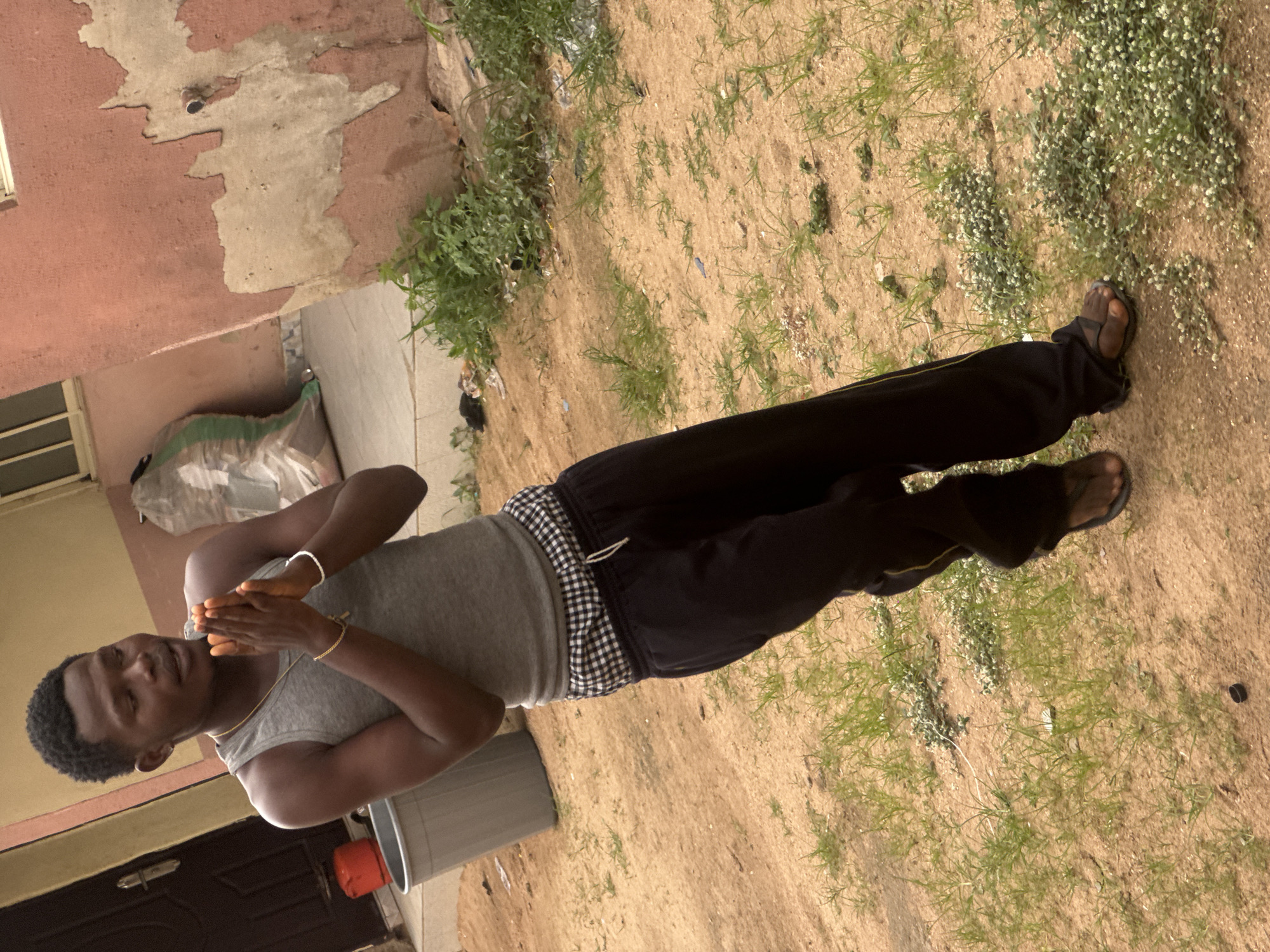 A young man with dark skin and short black hair is standing in what appears to be an outdoor area. He is wearing a gray tank top and black pants with a checkered waistband. His hands are clasped together in front of him, and he is looking towards the camera. He is also wearing a gold chain around his neck and a white bracelet on his left wrist.
In the background, there is a pink and beige building with peeling paint. To the left of the man, there is a doorway with a dark brown door. To the right of the man, there is a large gray trash can, and beyond that, a patch of sand with scattered green grass and small white flowers. The ground is a sandy color, suggesting a dry environment.
The lighting appears to be natural daylight, casting soft shadows. The overall impression is casual and candid. There is no visible text in the image. The man's expression is neutral.