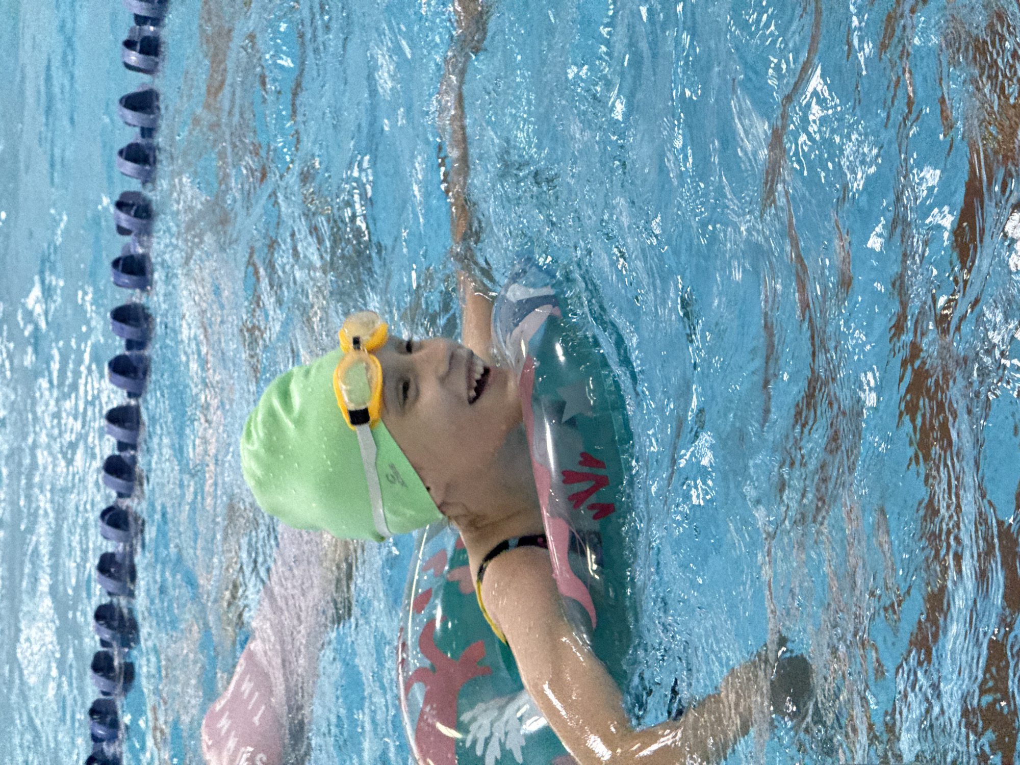 A young girl wearing a green swimming cap and yellow goggles swims in a pool. She is wearing a swimsuit and a floatation device around her waist. The pool has lane dividers. There are other swimmers in the background. The image was taken at the Olympic Swimming Pool in Birobidzhan, Russia.