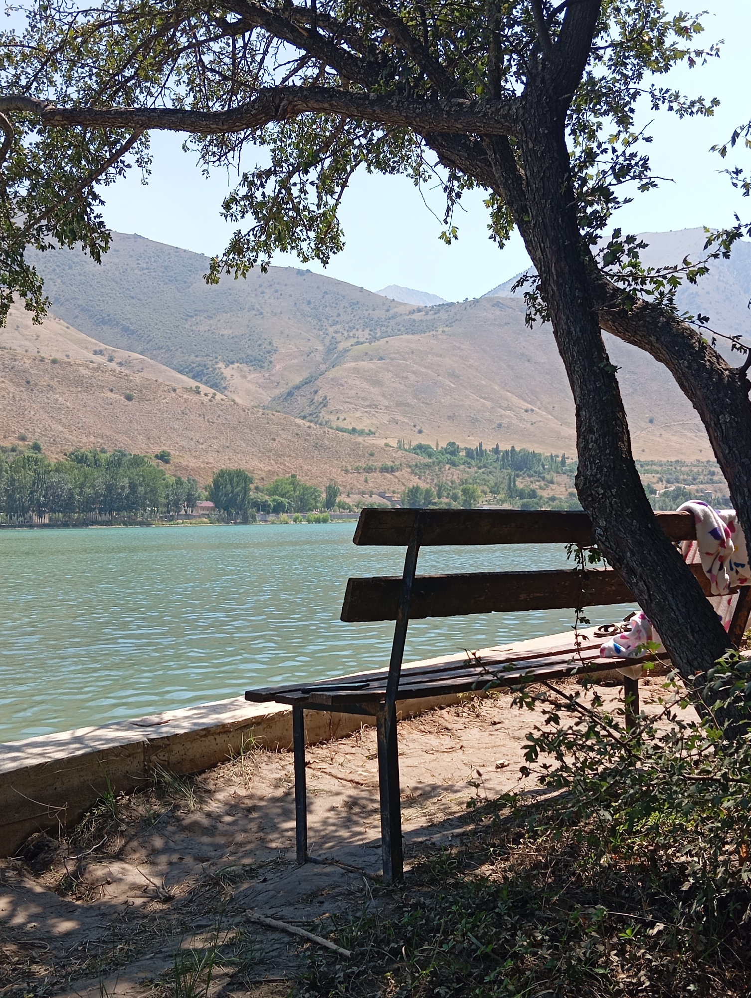 The image captures a serene lakeside scene, likely in the vicinity of Yukariyuz, Uzbekistan, judging by the arid, mountainous terrain and the presence of trees commonly found in the region. In the foreground, a weathered wooden bench is positioned under the dappled shade of a large tree. The bench is occupied by two folded blankets, one with a colorful floral pattern and the other with a lighter design. The lake's placid, green waters stretch out before the bench, its surface rippled by a gentle breeze. Along the lake's edge, a low concrete barrier separates the water from the sandy bank, which is dotted with patches of dry grass and fallen leaves.
Beyond the lake, a dense line of trees forms a verdant border, with a small building nestled amongst them, hinting at a settlement or a park area. The background is dominated by undulating, dry hills that rise towards a clear, pale blue sky. The overall impression is one of tranquility and natural beauty, a peaceful spot for relaxation by the water.
