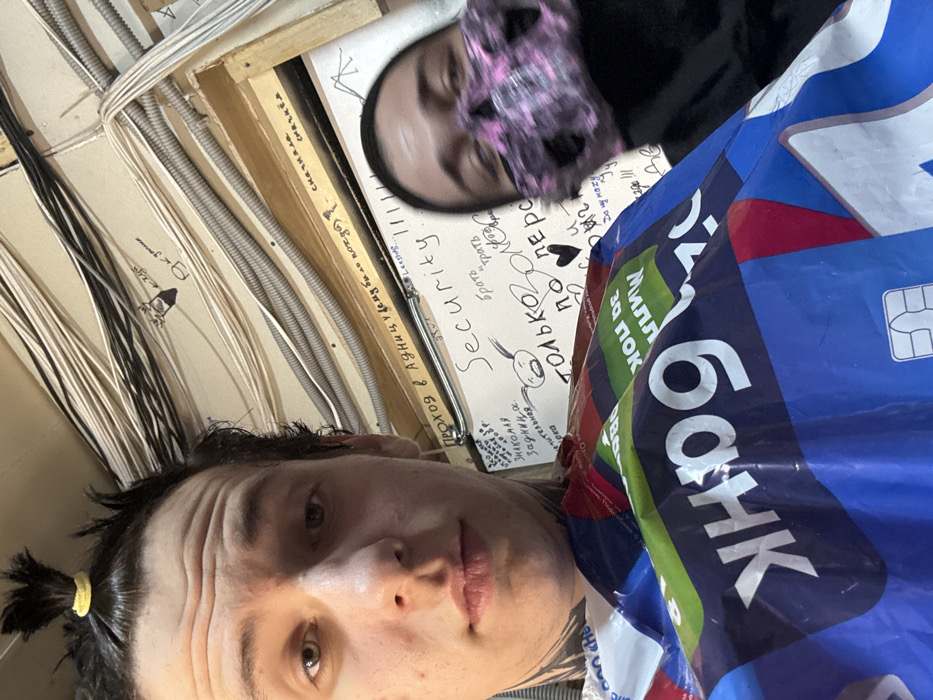 Two people are in the image. One person in the foreground has their hair in a ponytail. In the background, the second person is wearing a face covering. The surroundings include an electrical system, a blue bag with white letters spelling "банк", and handwritten notes on a surface that mention "Security" and "только оло по перс". The scene appears to be indoors.