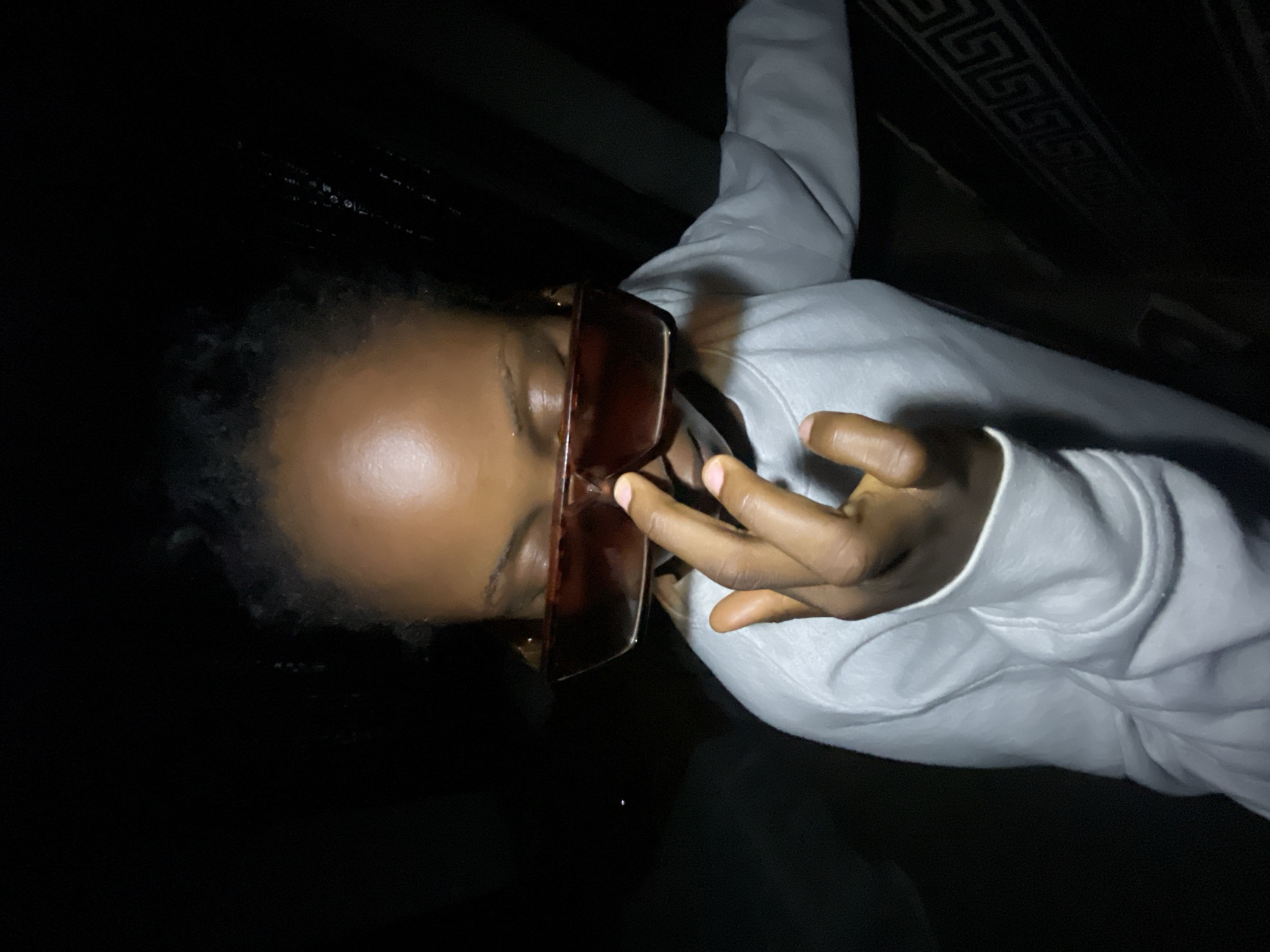 A young Black person, likely a teenager, is photographed up close in low light conditions. They are wearing a light-colored sweatshirt and large, dark, angular sunglasses that cover their eyes. Their right hand is raised, with fingers touching the bridge of the sunglasses and the tip of their nose. The person's face is angled slightly downward, and their gaze is obscured by the sunglasses. The background is dark and out of focus, with hints of a dark, possibly upholstered surface with a geometric pattern visible on the right side. There are no discernible urban features or specific landmarks of Lokoja, Nigeria, visible in the image. The lighting appears to be artificial and directional, creating strong shadows and highlighting the subject's face and hand. The overall impression is an intimate, candid shot, possibly taken indoors or in a dimly lit outdoor setting.