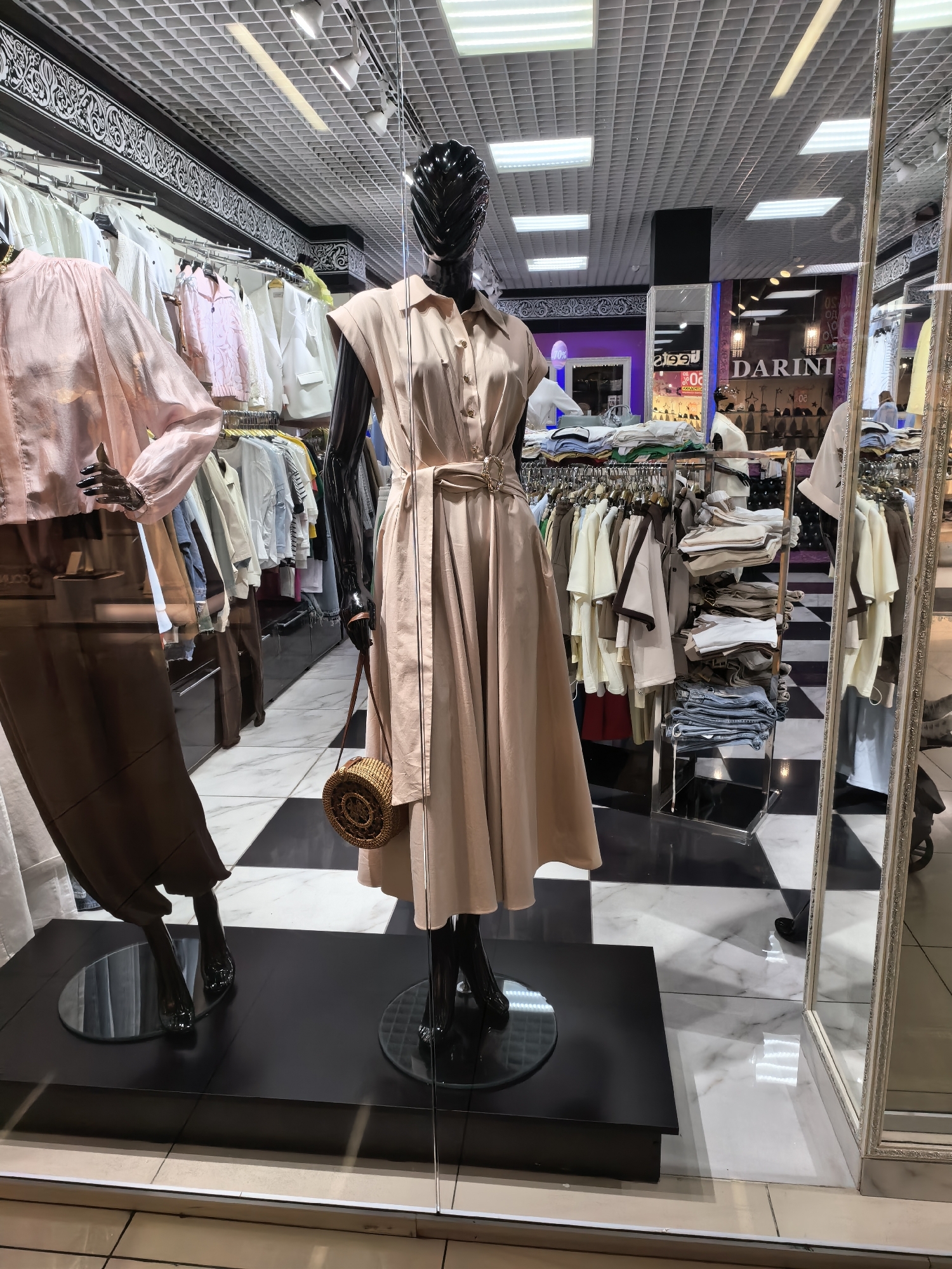 The image shows a retail store window display featuring two mannequins dressed in women's clothing. The mannequin on the right is wearing a light pink, collared, sleeveless midi dress cinched with a fabric belt, and carrying a small, round, woven straw bag. The mannequin on the left is wearing a pale pink, loose-fitting blouse and a long, dark brown skirt. Behind the mannequins, racks of clothing are visible, displaying various garments such as shirts, jackets, and folded stacks of fabric. The store interior features a black and white checkered floor pattern and white marbled flooring. A large decorative mirror with an ornate frame is positioned to the right of the display. The store is identified as "DARINI" based on signage visible in the reflection. The scene is likely a clothing boutique in the city of Sterlitamak, Russia.