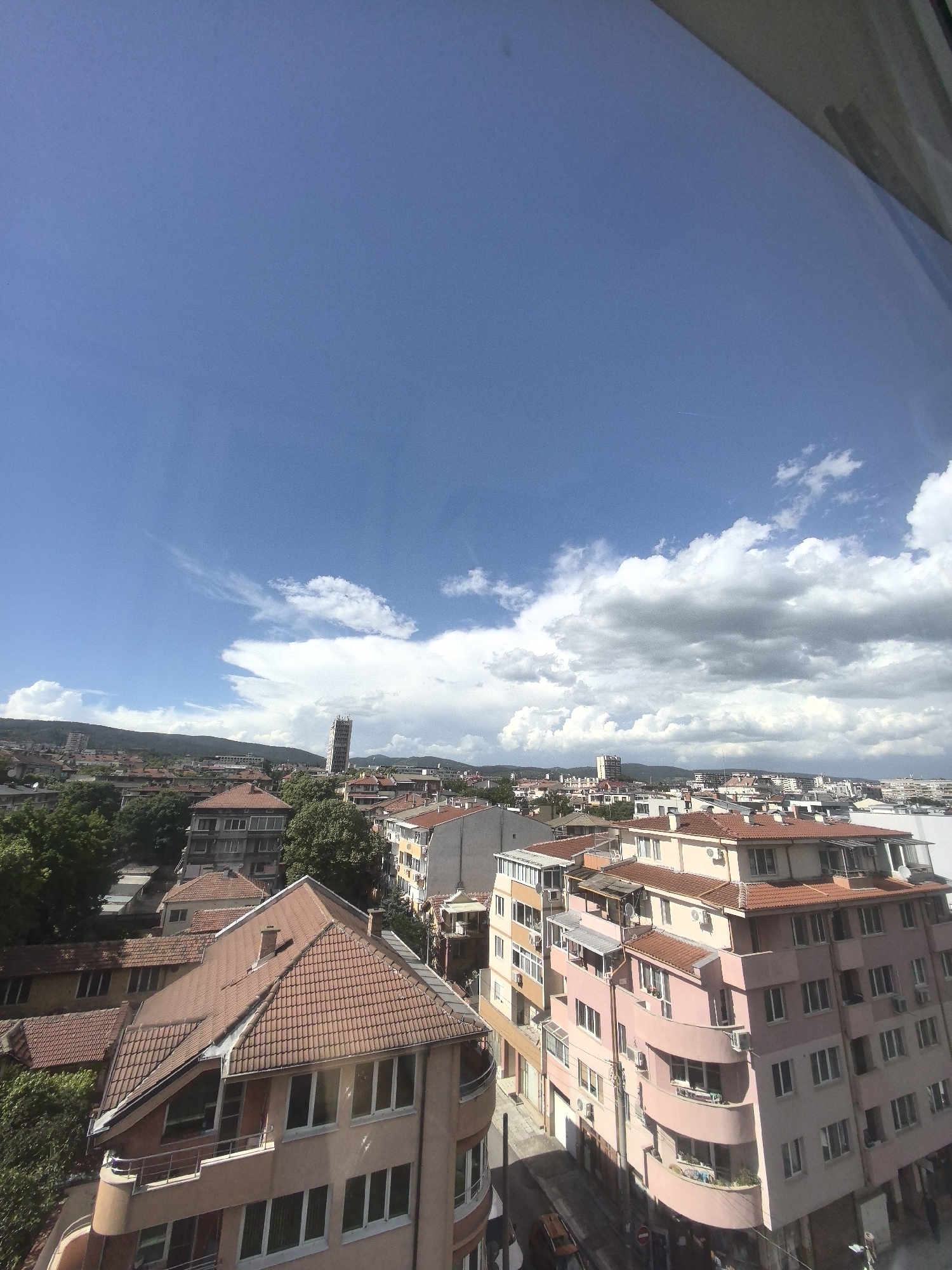 The image shows an urban landscape in the city of Unknown, Unknown. The view is from a high vantage point, possibly a window. The scene includes buildings with terracotta roofs, green trees, and a blue sky with some white and gray clouds. In the background, there are mountains or hills. A tall tower-like structure can be seen in the distance. The nearest building has a sloping, red-tiled roof and is multi-story. A pinkish building with curved balconies is also prominent in the view. A street runs between the buildings, suggesting a residential or commercial area. The sky is mostly clear with scattered clouds. The angle and composition of the image suggest it was taken looking down on the city.