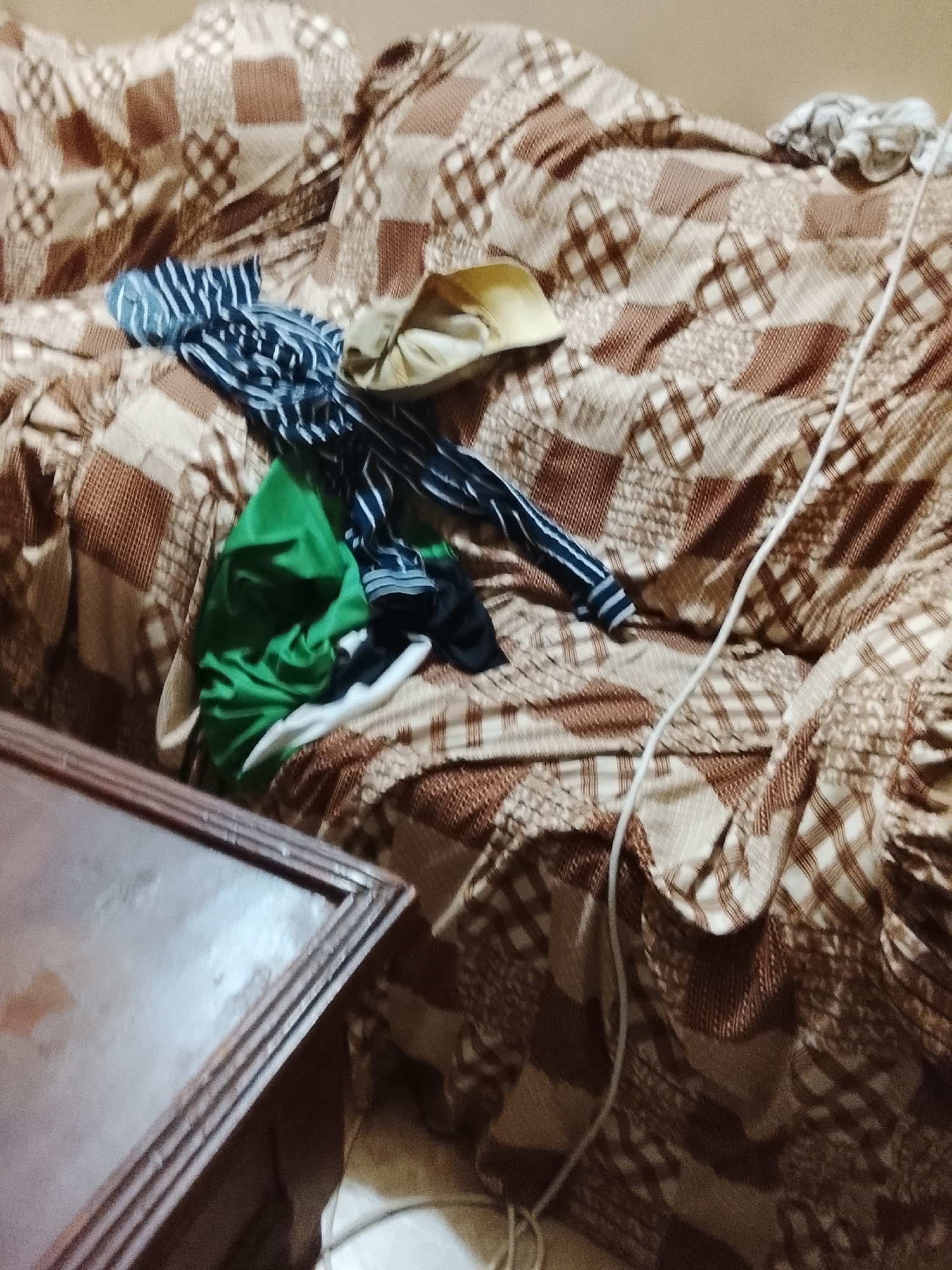 The scene depicts a disheveled couch with a brown and beige patchwork pattern cover. Scattered on the couch are various items of clothing, including a green shirt, a striped blue and white shirt, a yellow hat, and some other pieces of unidentified garments. A white cable is draped across the couch. A portion of a wooden table is visible in the lower-left corner.
There is no contextual information in the image that suggests that this location is in Multan, Pakistan.