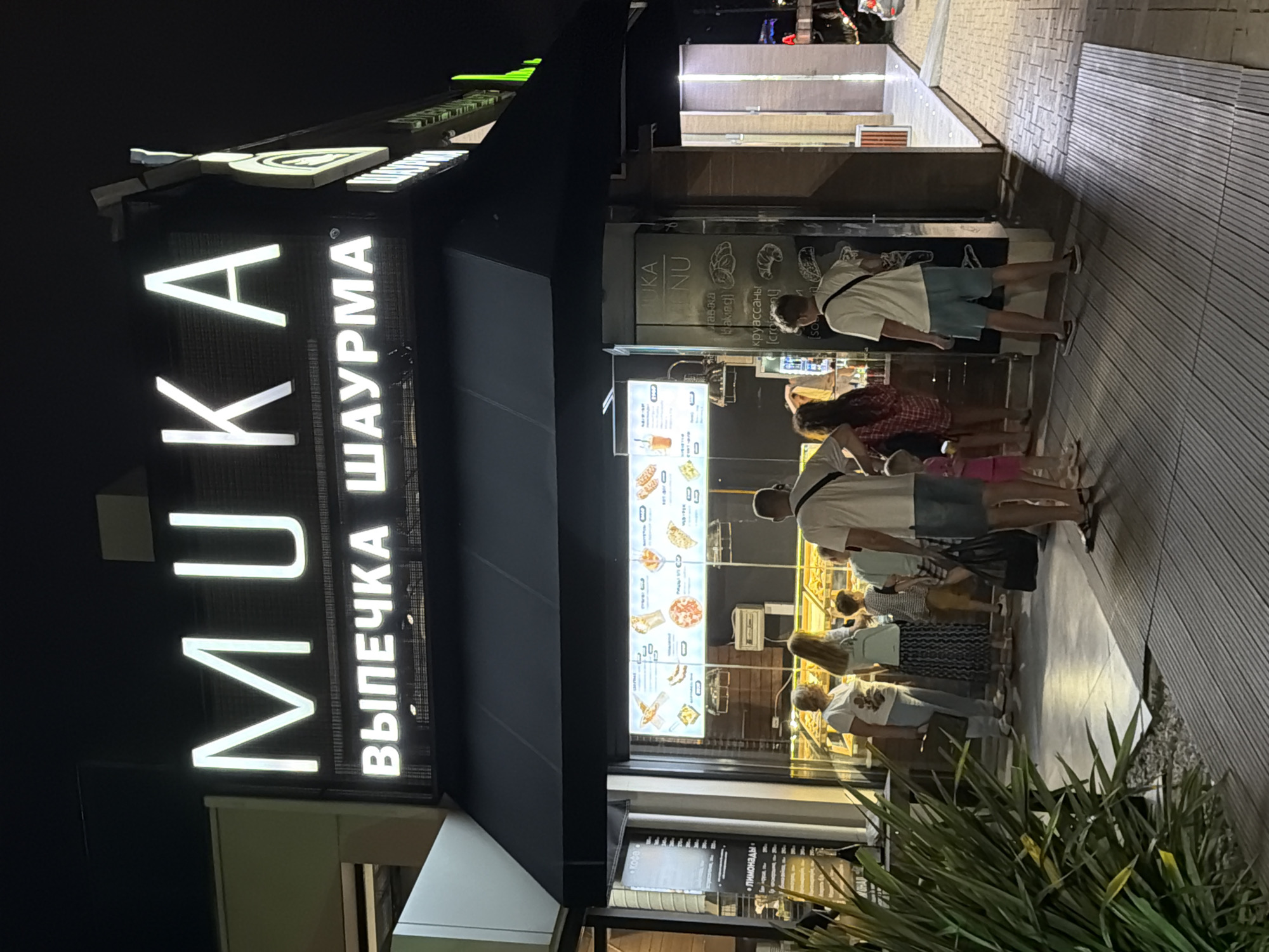 This is a nighttime outdoor shot of a food establishment in Sochi, Russia. The primary subject is a storefront with illuminated signage, displaying the words "MUKA" and "ВЫПЕЧКА ШАУРМА" (Bakery Shawarma). A menu board is also visible, featuring images of various food items like pizzas, croissants, and what appear to be savory pastries.
Several people are standing in line outside the establishment, seemingly waiting to order or pick up food. They are dressed in casual summer attire, suggesting warm weather. The atmosphere appears relaxed and ordinary, with no specific event being depicted beyond the daily operations of the food vendor. The ground is paved with wooden planks, and there are decorative plants in the foreground. The lighting from the signs and the interior of the establishment creates a warm and inviting ambiance, contrasting with the dark night sky. The presence of Cyrillic text on the signage and menu confirms the Russian location.