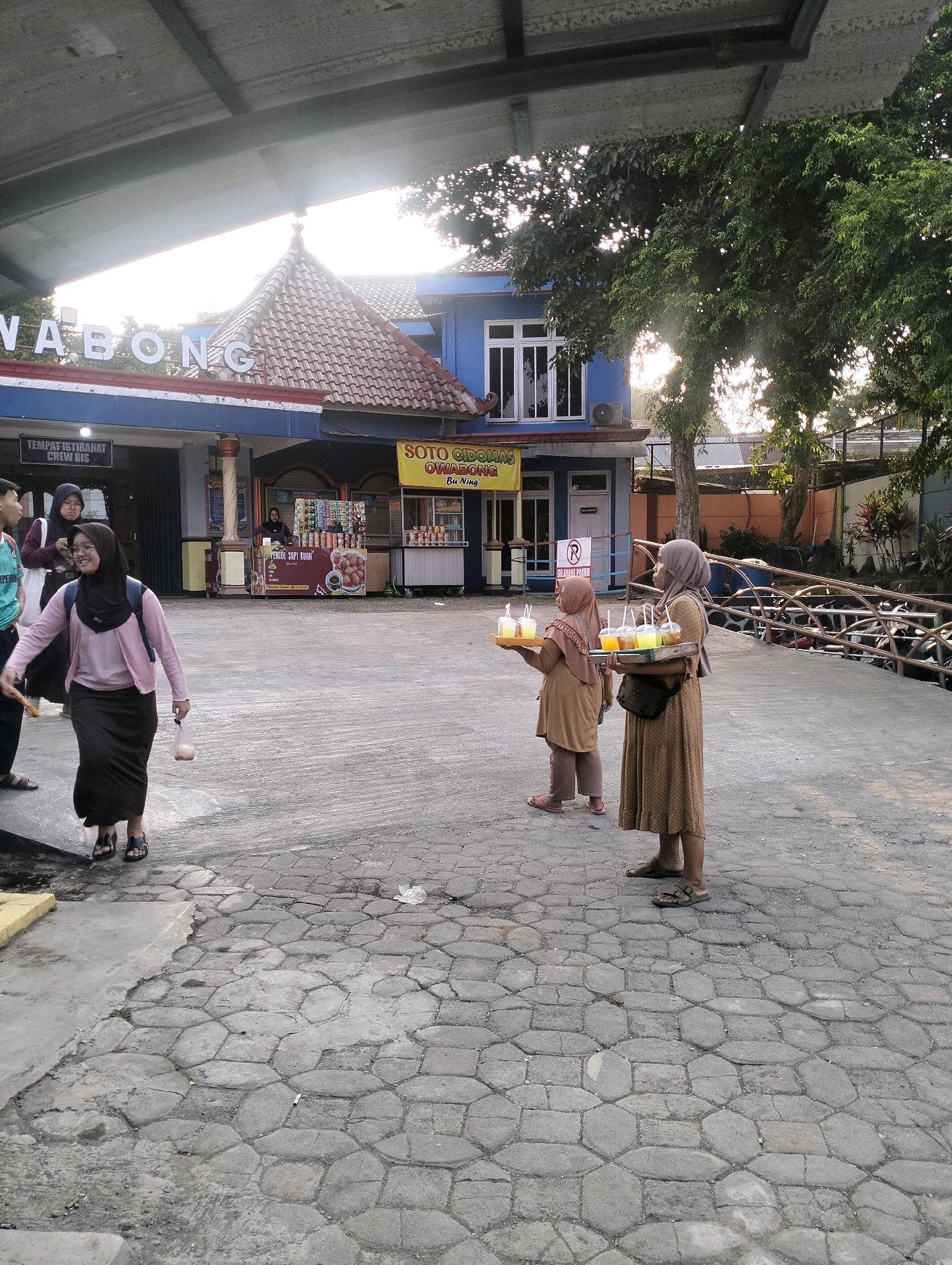 The image depicts the exterior of a roadside establishment, likely a food and drink stand, in Karangbanjar, Indonesia. The establishment has a blue facade with a distinctive red-tiled roof, and a sign that appears to read "OWABONG". In the foreground, a woman wearing a pink cardigan over a long brown skirt and hijab walks away from the camera, holding a plastic bag. To her right, two women are standing, each carrying a tray laden with plastic cups filled with brightly colored beverages. The ground is paved with hexagonal stones. In the background, there are trees, a metal fence, and other buildings. The overall atmosphere suggests a warm, possibly late afternoon, setting.