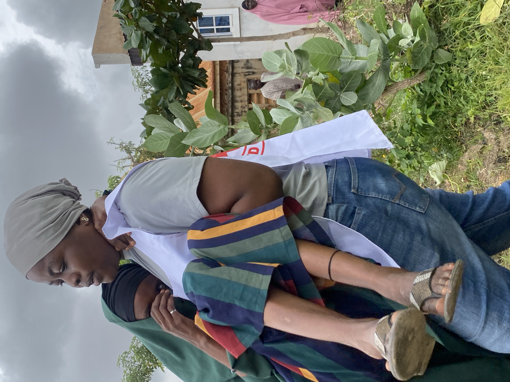 In Kawo, Nigeria, a woman in a grey headscarf carries another person, who is wearing a green hijab and appears distressed, with their face buried in the carrier's shoulder. The carrier is wearing a white sash with red lettering and blue jeans. The person being carried is wearing a colorful striped garment. The background shows a building and green vegetation. Two other people are visible in the distance, one in a light-colored garment and another in a pink garment. The sky is overcast and grey.