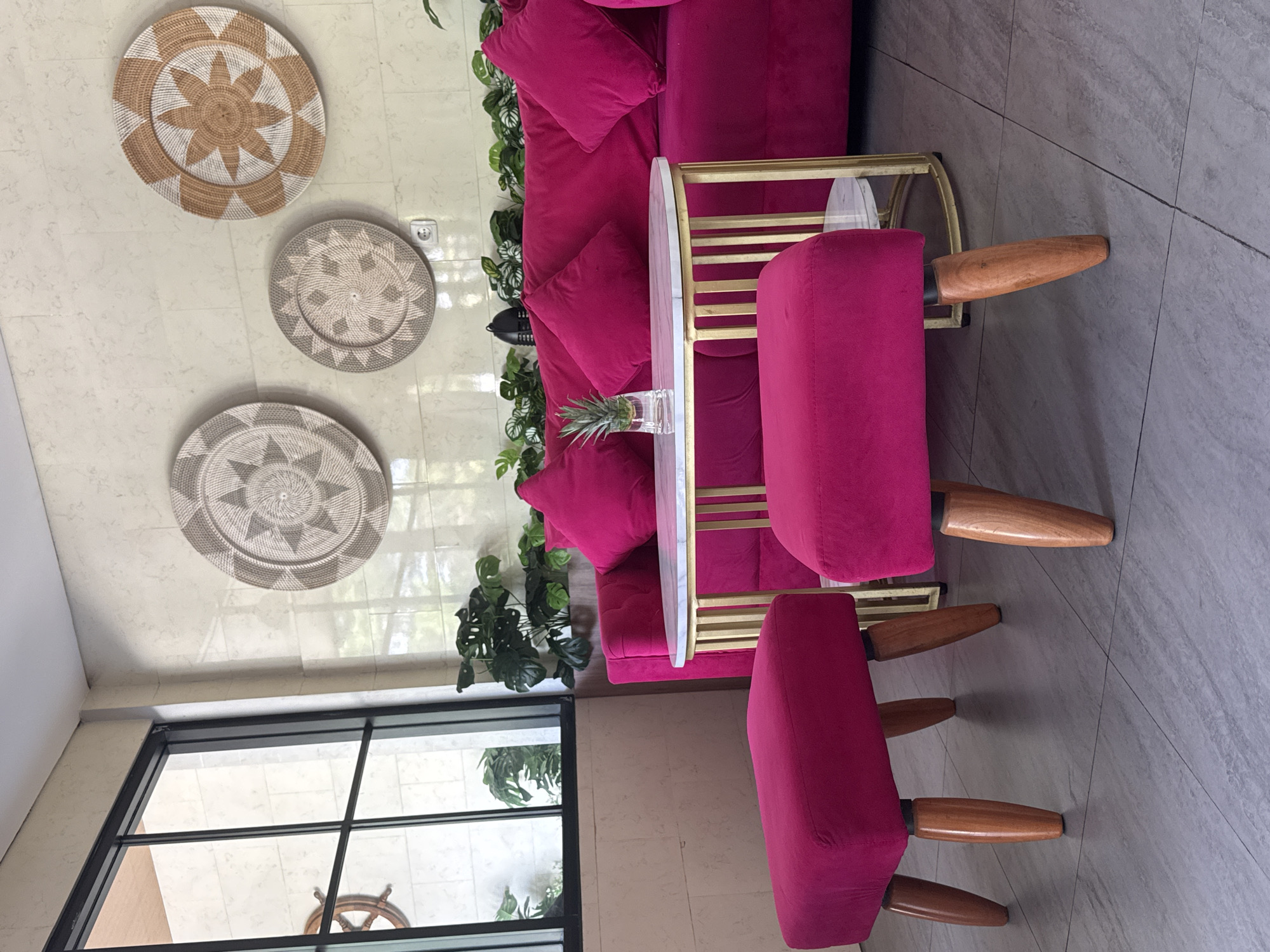 The image captures an interior setting, potentially within a hotel lobby in Kuta, Indonesia, given the style and furnishings. The focal point is a magenta-colored sofa with matching chairs arranged around a small, oval-shaped side table. The table has a white marble-like top and a gold-colored frame. A pineapple in a clear glass vase sits on the table.
The wall behind the sofa is decorated with three decorative woven wall hangings. To the left, a large window offers a view of greenery outside, possibly a garden or landscaped area. The flooring appears to be tiled in a light gray color, contributing to a modern aesthetic. No people are visible in the image.
