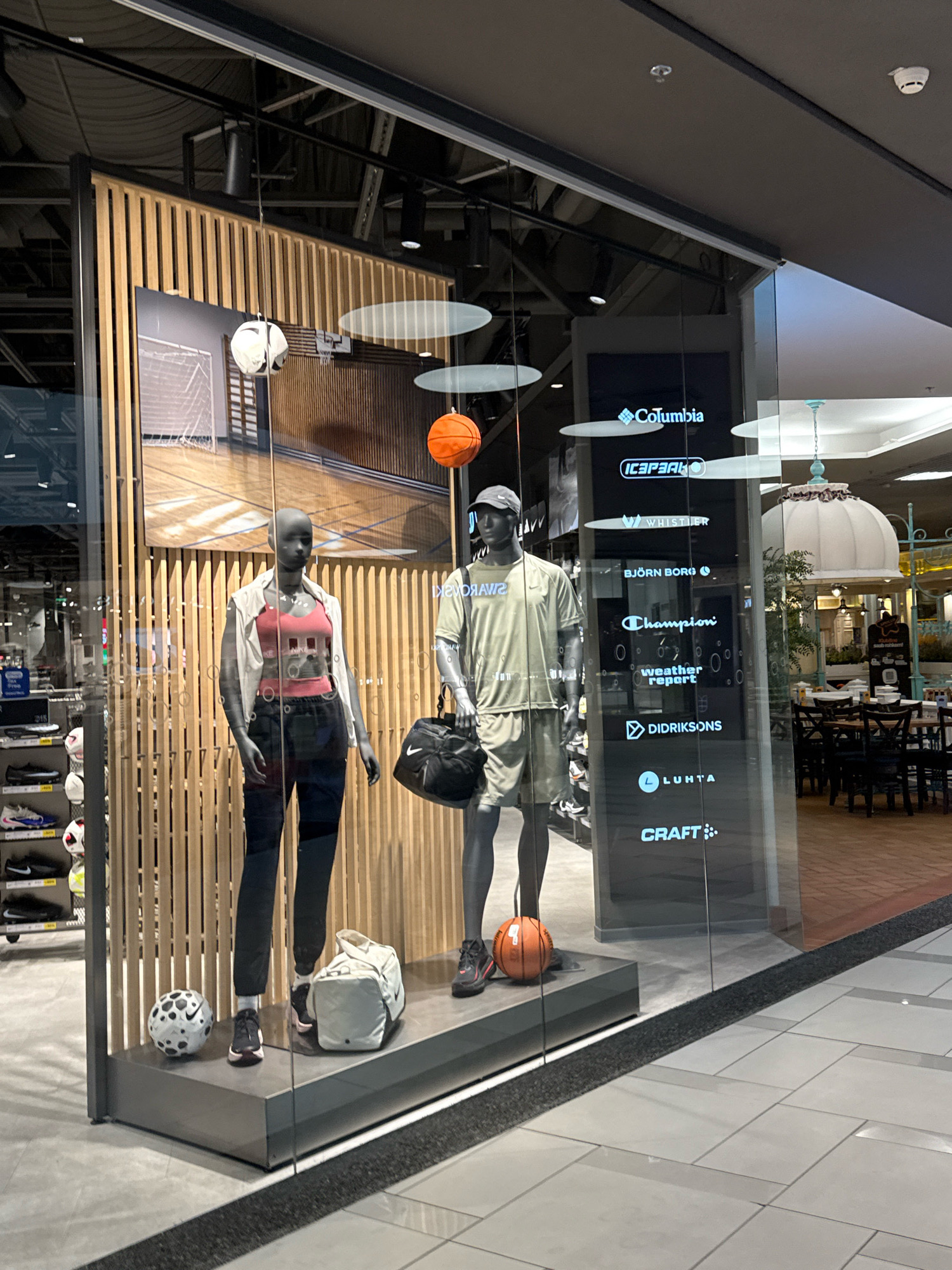 This is an eye-level, medium shot of a retail store window display featuring two mannequins dressed in athletic wear. The display is in a shopping mall, possibly in Tallinn, Estonia, as suggested by the visible brand logos on a vertical digital sign.
The left mannequin is female, wearing a white sleeveless vest over a red tank top with a black Nike logo, and dark pants. A white duffel bag sits at her feet next to a black and white patterned soccer ball.
The right mannequin is male, wearing a light green t-shirt and shorts, with a grey baseball cap. He is holding a black duffel bag and standing next to an orange basketball.
Behind the mannequins, a wooden slatted wall forms the backdrop. Above them, white oval shapes hang suspended, and an image of a basketball court is visible on a screen.
The digital sign to the right displays several brand logos, including Columbia, Icepeak, Whistler, Björn Borg, Champion, Weather Report, Didriksons, Luhta, and Craft.
The foreground shows the tiled floor of the shopping mall and a dark rubber matting at the edge of the display. Through the glass, the interior of the mall is visible, with seating areas, tables, and chairs in a restaurant or cafe.