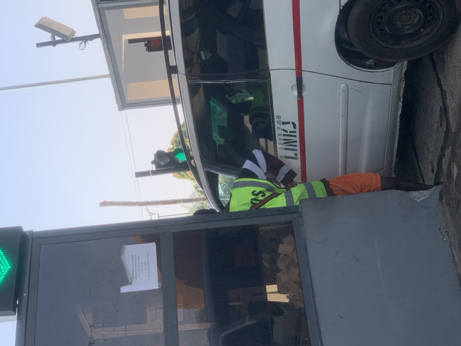 This image depicts a checkpoint or toll plaza in Kubare, Nigeria. A male attendant is positioned between a dark-paneled booth structure and the driver's side of a white multi-purpose vehicle. The attendant wears a neon green reflective vest with "POS" visible on the back, orange shorts, and a long-sleeved shirt with black and white striped sleeves; his right arm is bent, extended towards the vehicle's interior. The white vehicle, possibly a minivan, displays "LINKS" in black capital letters and a horizontal red stripe on its side. Its driver's side front window is partially open.
Above the scene, a large green traffic light indicates an open lane, while a smaller red traffic light is visible further down the lane, above the vehicle. A white rectangular sensor or antenna is mounted on a pole above the vehicle. Taped to the interior of the booth's dark glass window is a white sign with black text, partially visible, stating: "CASH PAYMENT NOT ALLOWED. PAYMENT METHOD INCLUDED. DON'T CREATE TRAFFIC. TRANSIT. THIS LANE IS FOR ELECTRONIC PAYMENT." The sky is clear, and a utility pole stands in the background.