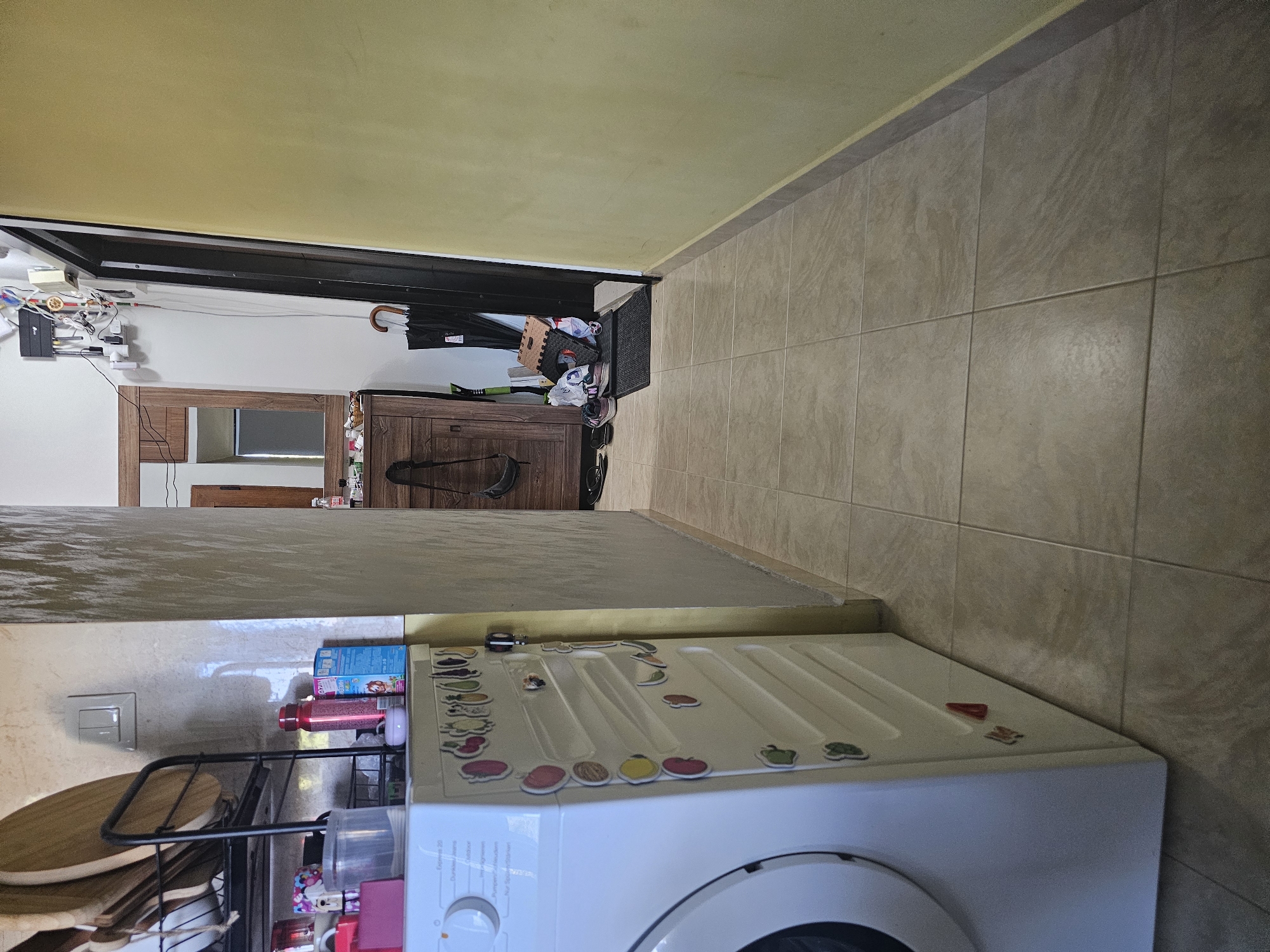 The image depicts an interior hallway. On the left, a white washing machine is partially visible, adorned with numerous colorful fruit-shaped magnets on its side. To the left of the washing machine is a black dish rack holding various plates. In the background, a wooden cabinet with drawers and a mirror above it stands against a beige wall. Next to the cabinet, a black umbrella is propped upright, and various bags and shoes are scattered near a dark doorway. The floor is covered with beige square tiles. The right wall is painted a pale yellow. The scene appears to be located in a residential setting, possibly a kitchen or laundry area within an apartment in Sofia, Bulgaria.