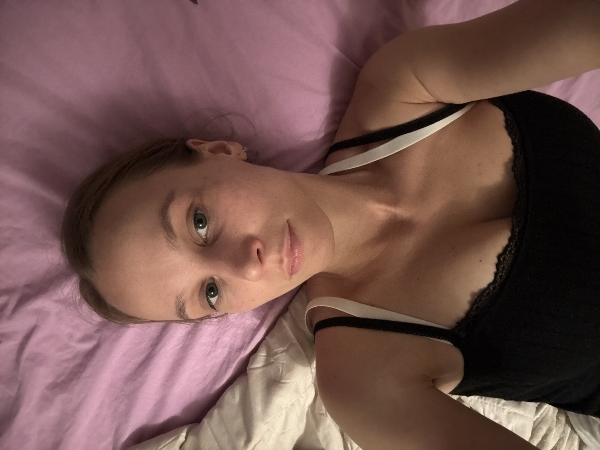 A young woman is lying in bed, looking up at the camera. She has fair skin, green eyes, and her hair is pulled back. She is wearing a black tank top with white straps. The bed is covered with a pink comforter and a white duvet. The image appears to have been taken indoors in a bedroom. There is no indication of the specific location within Überherrn, Germany.