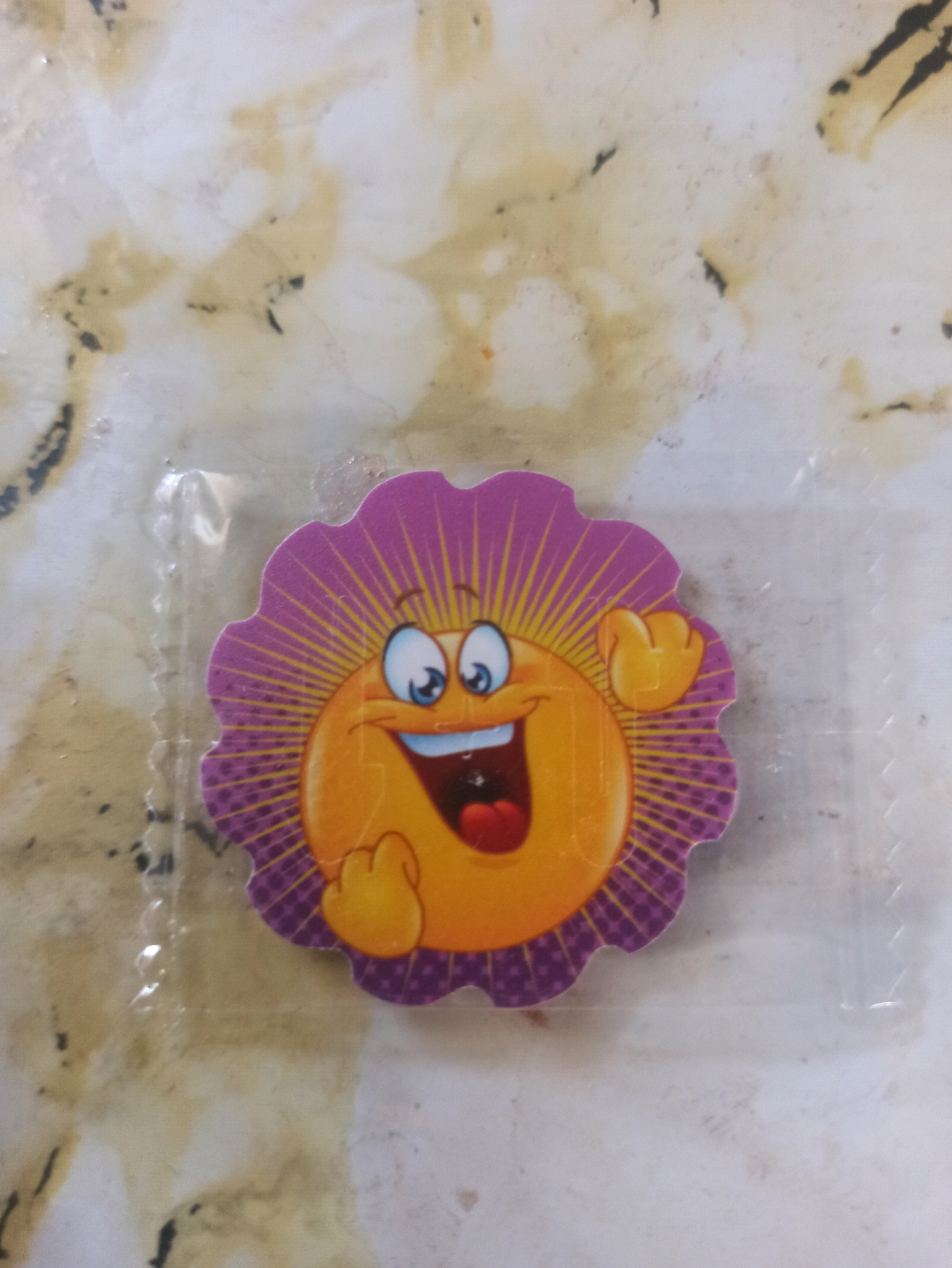 An image shows a sticker with a sun-like cartoon character. The character has a smiling face with big blue eyes, a wide mouth open in a grin, and a red tongue. It has orange "rays" radiating from its head, with a purple, scalloped border. One yellow, cartoonish hand is raised in the air. The sticker appears to be in plastic packaging, and is lying on a surface with a mottled, light-colored pattern. No specific location within Machraa El Ain, Morocco is discernible from the image.