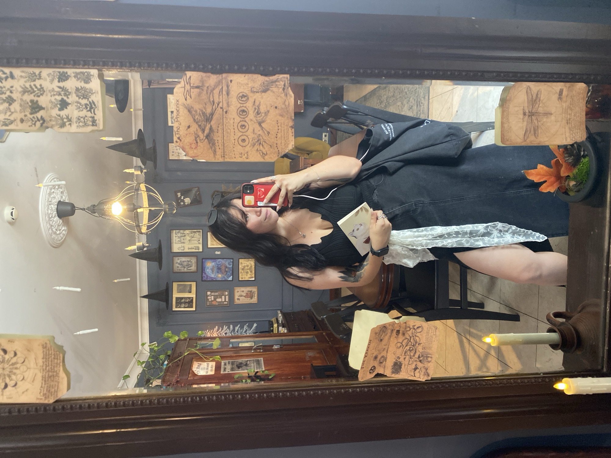 A young woman with dark hair is taking a selfie in a mirror in what appears to be a room decorated in a witchy or gothic theme. The room has dark blue walls, and is adorned with several framed pictures of botanical drawings, moon phases, and birds. There are also black witch hats hanging from the ceiling, and faux floating candles. In the foreground of the mirror, two battery-operated candles are lit. The woman is wearing a black ribbed tank top with a plunging neckline and a long black denim skirt with a high slit. She is holding a small book in her left hand and her smartphone in her right hand, with earbuds in her ears. A white lace scarf is draped over her left arm. In the reflection, a dining table with chairs can be seen, along with a wooden cabinet filled with items. The overall impression is of a quirky and themed interior, possibly a shop or a private residence with a specific aesthetic. The location is identified as Saint Petersburg, Russia, based on the provided prompt.
