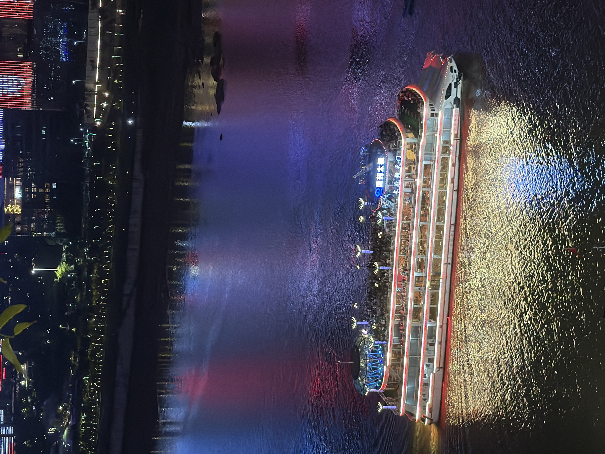 An aerial, medium shot captures a brightly lit, multi-decked passenger boat navigating a dark body of water at night. The boat, adorned with red and white lights, has a large crowd of people visible on its upper decks. The water's surface reflects the boat's illumination and the vibrant colored lights from the distant city skyline, creating a shimmering, colorful spectacle. The city, illuminated by numerous lights, forms a backdrop of buildings and streetlights along the waterfront. The scene is likely in Yuzhong District, China, given the context of illuminated cityscapes and large passenger boats often seen on rivers in such urban areas.