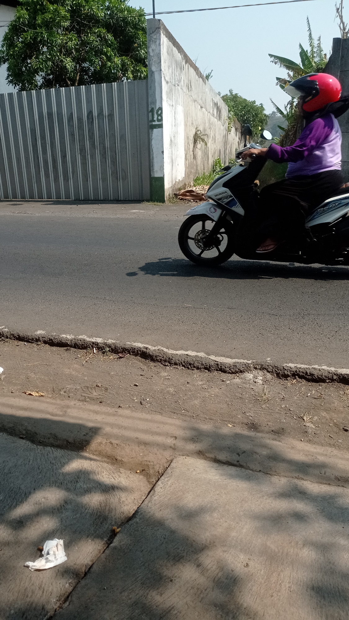 A person wearing a red helmet and a purple long-sleeved shirt is riding a white and black motorcycle on a paved road. The motorcycle has the brand name "HONDA" visible. The person is positioned in the right half of the image, and the motorcycle is facing left. In the background, there is a tall, light-colored concrete wall with vertical metal siding to its left. The number "18" is painted in green on the concrete wall. To the right of the concrete wall, there are some banana trees. The road is bordered by a raised dirt and gravel area. In the foreground, there is a concrete surface with shadows cast by trees and foliage. A crumpled white piece of paper is visible on the concrete surface in the lower left corner of the image. The sky is clear and blue.