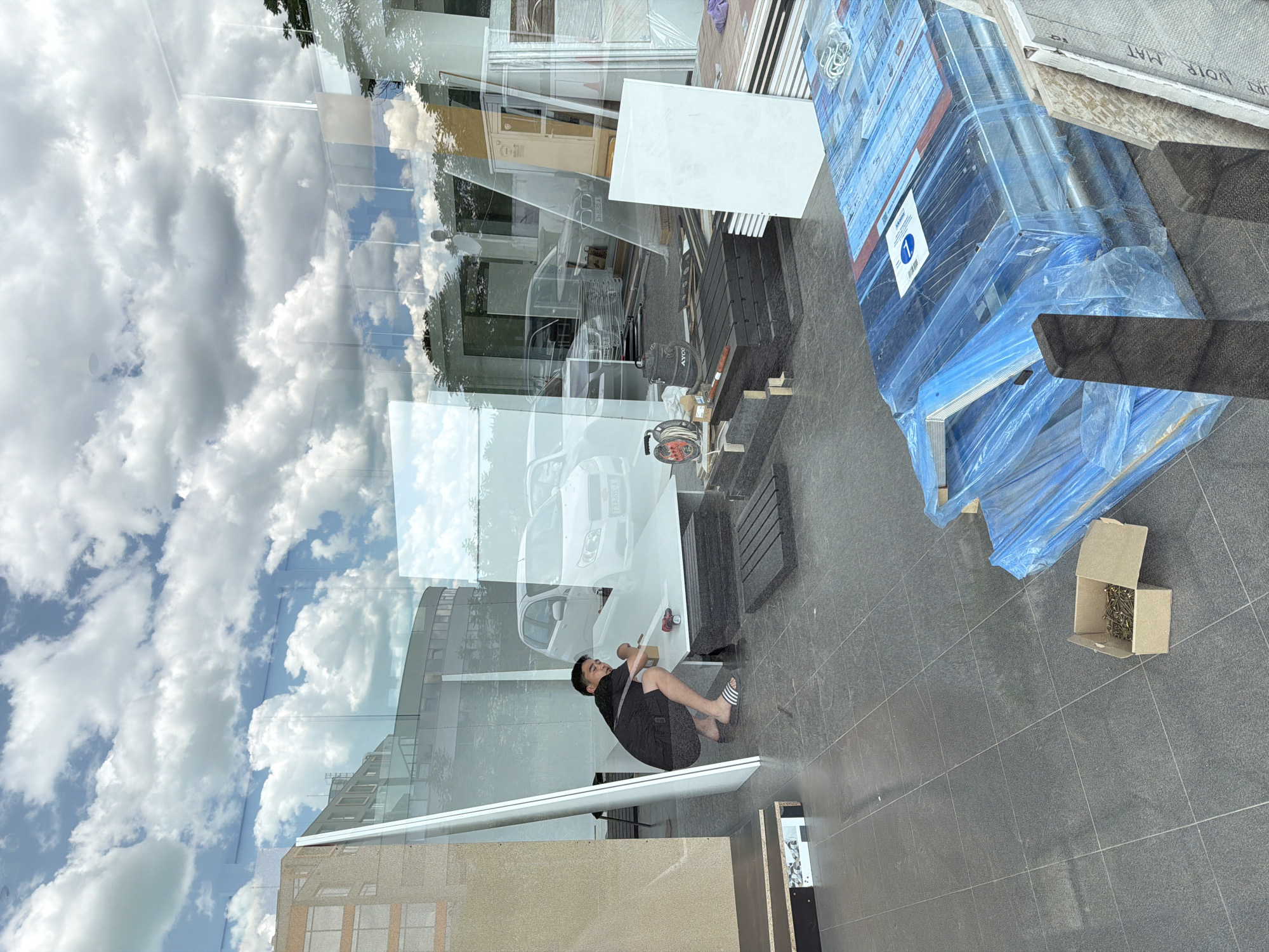 The image shows a man squatting on a tiled surface, next to a cardboard box containing fasteners. The man is wearing a black shirt, dark shorts, and black and white striped sandals. Behind the man are building materials including wood and metal, some of which are wrapped in blue plastic. A cloudscape is visible through a large glass window. Reflected in the glass are buildings. The reflection is a modern building. The location is Burgas, Bulgaria.